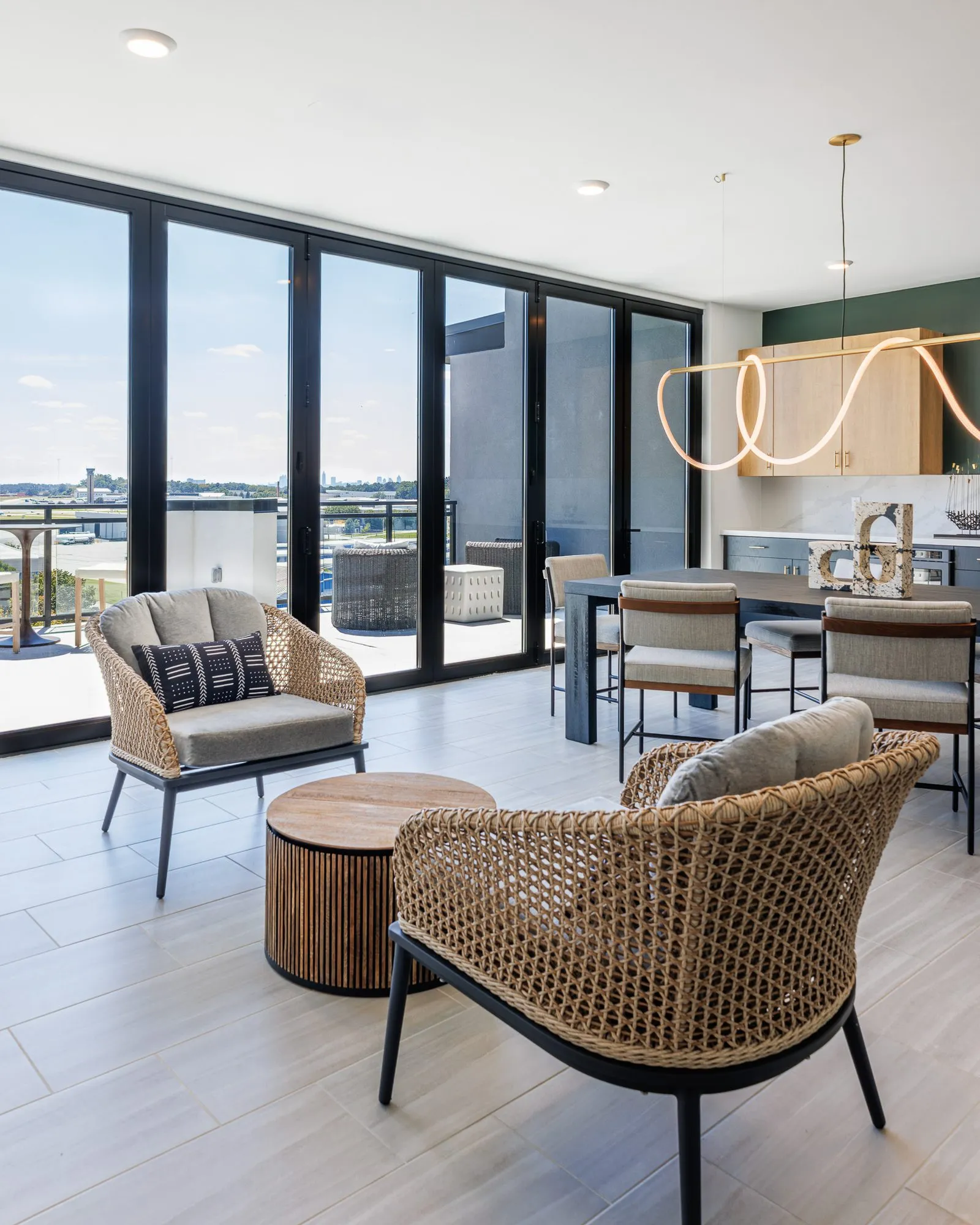 Multifamily amenity lounge with woven armchairs and a fluted drum table by the folding glass wall, rope light over the table