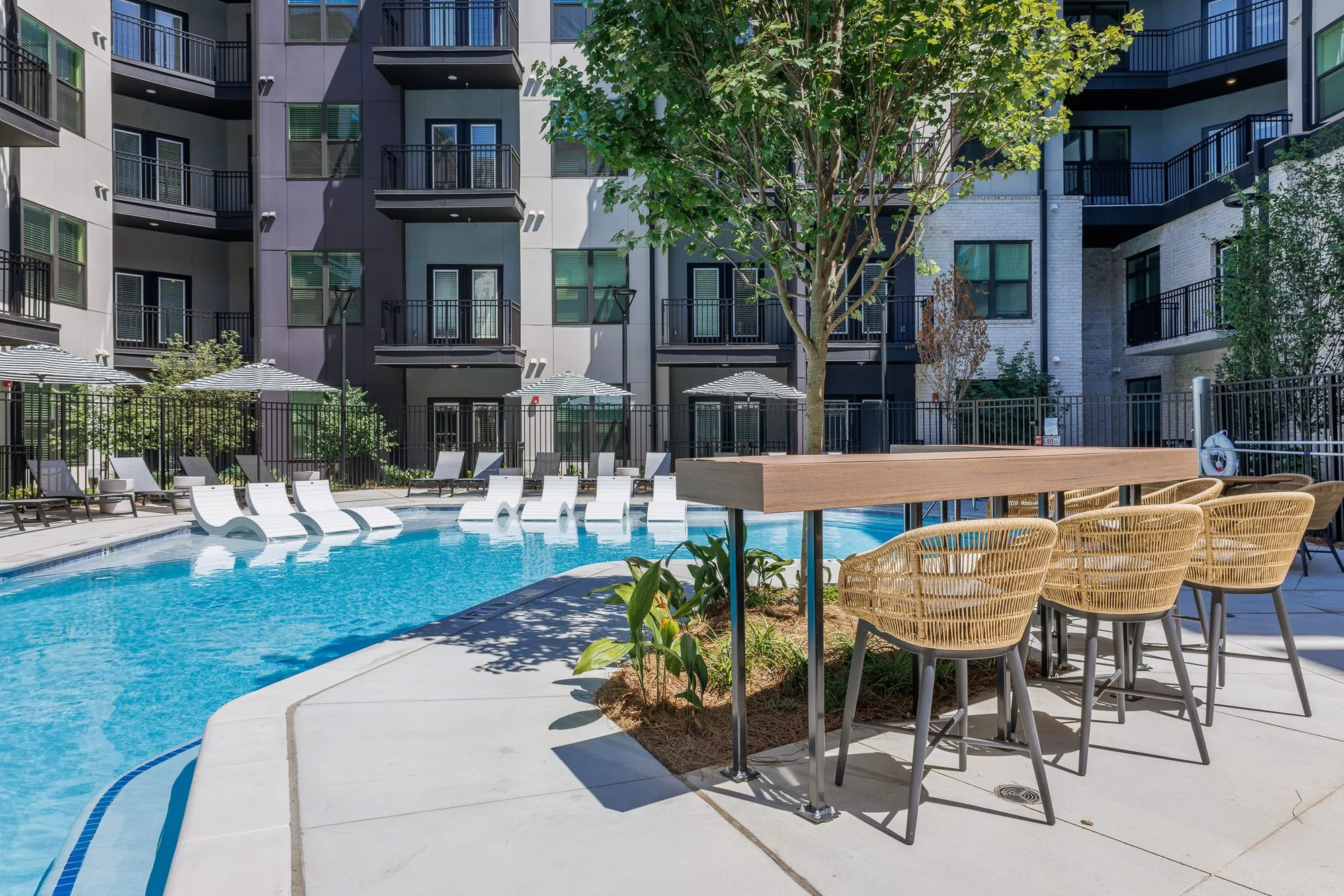 Multifamily amenity pool with white chaises on the sun shelf, woven counter stools at a high wood table, residences around