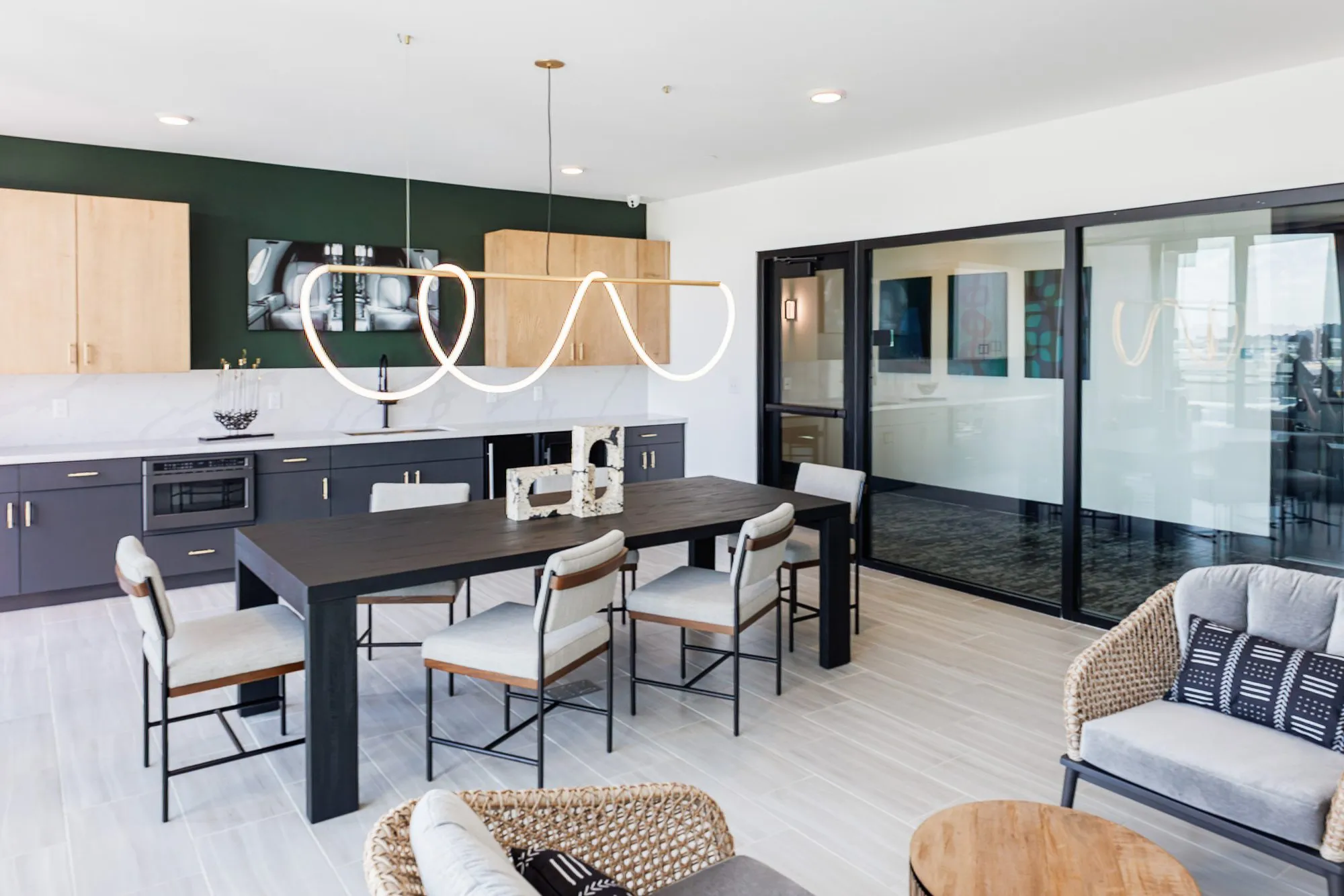 Multifamily amenity clubroom with a dining table and grey chairs under a looping rope light, woven lounge chairs in front