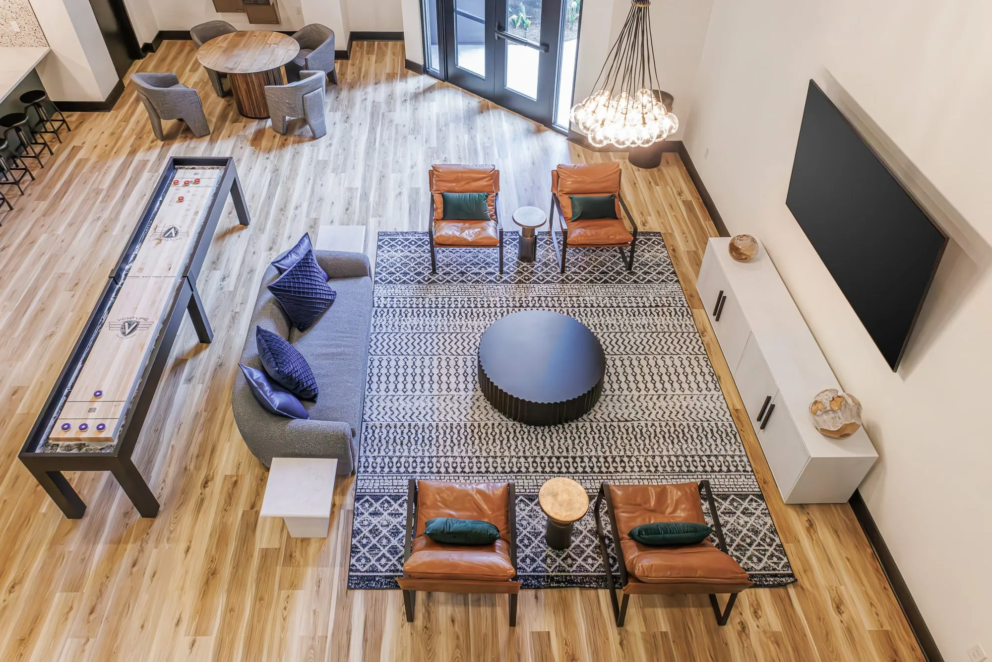 Multifamily amenity clubroom from above, leather armchairs around a black drum table on a geometric rug, shuffleboard beside
