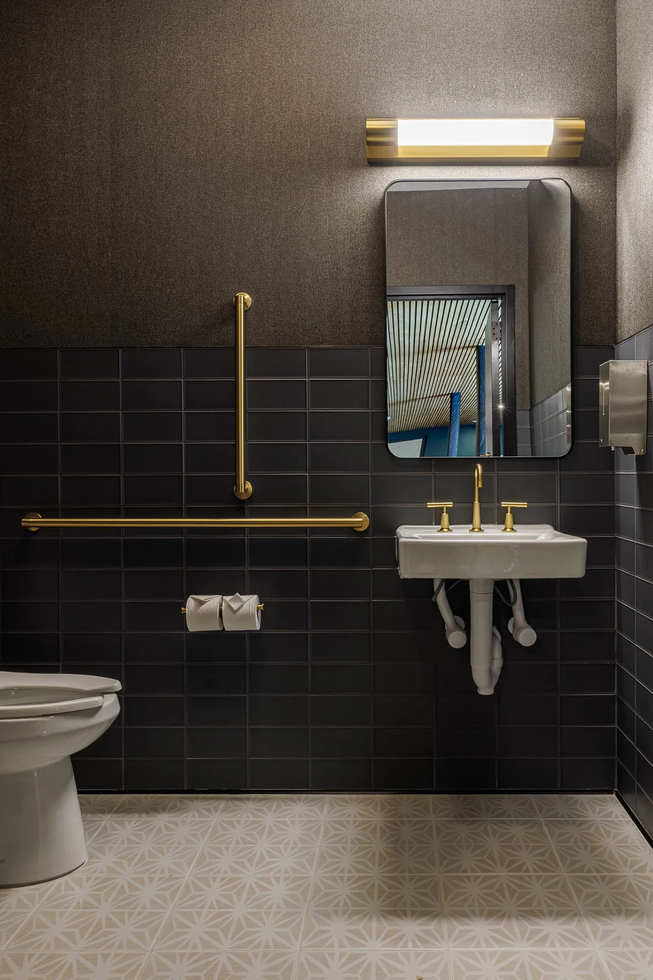 Multifamily amenity restroom with charcoal tile, a wall-hung basin with brass taps and grab bars under a brass bar light