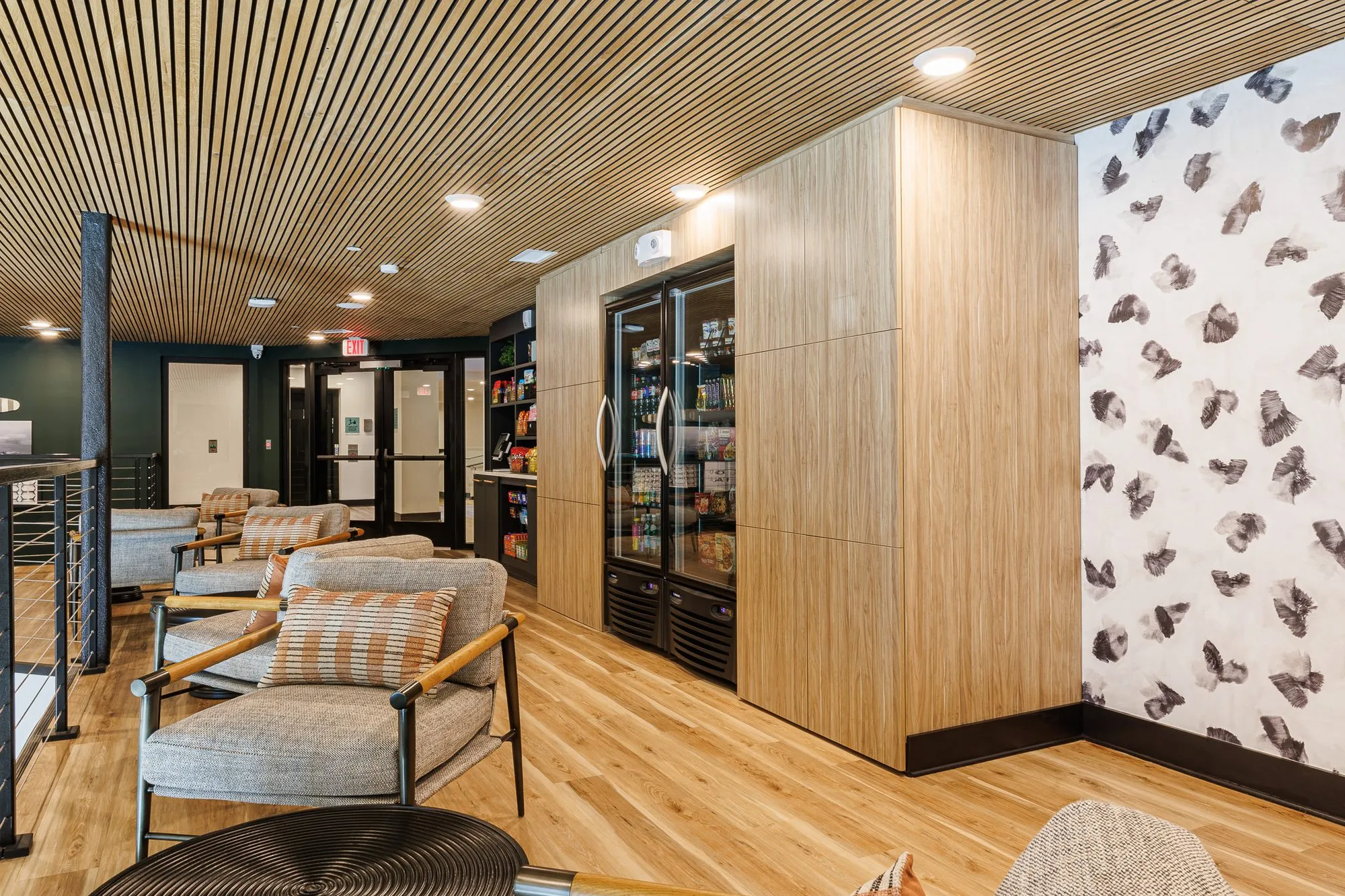 Multifamily amenity lounge with grey armchairs and a market fridge set into oak panelling beside butterfly wallpaper