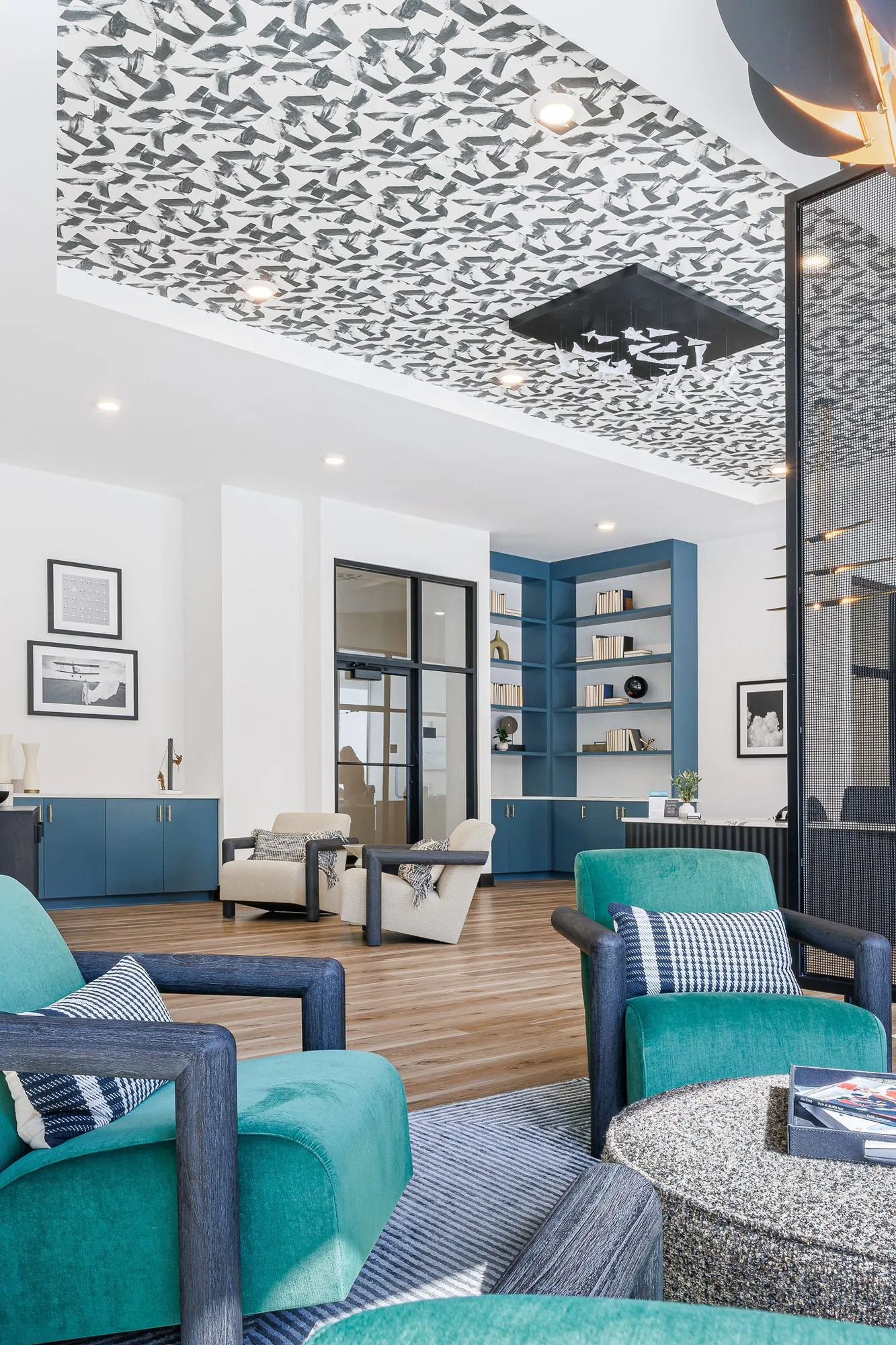Multifamily amenity lounge with teal armchairs and a speckled ottoman under a patterned ceiling panel, blue shelving beyond