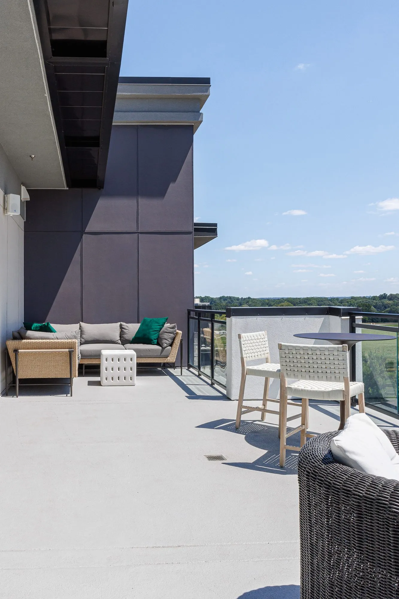 Multifamily amenity rooftop terrace with a woven sofa and white counter chairs at a high table by the glass rail