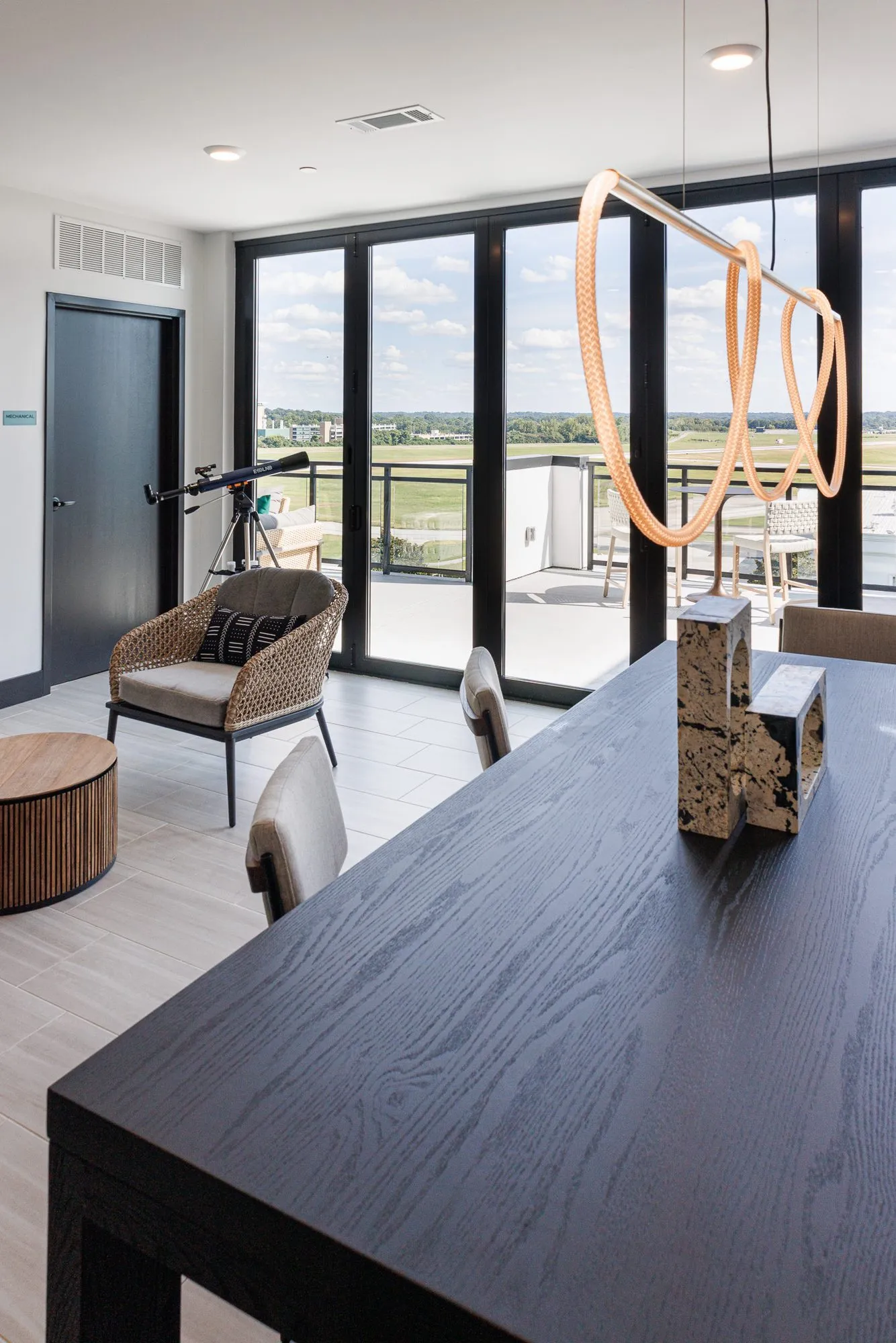 Multifamily amenity clubroom with a telescope on a tripod by folding glass doors, a woven chair and dark table, a rope light