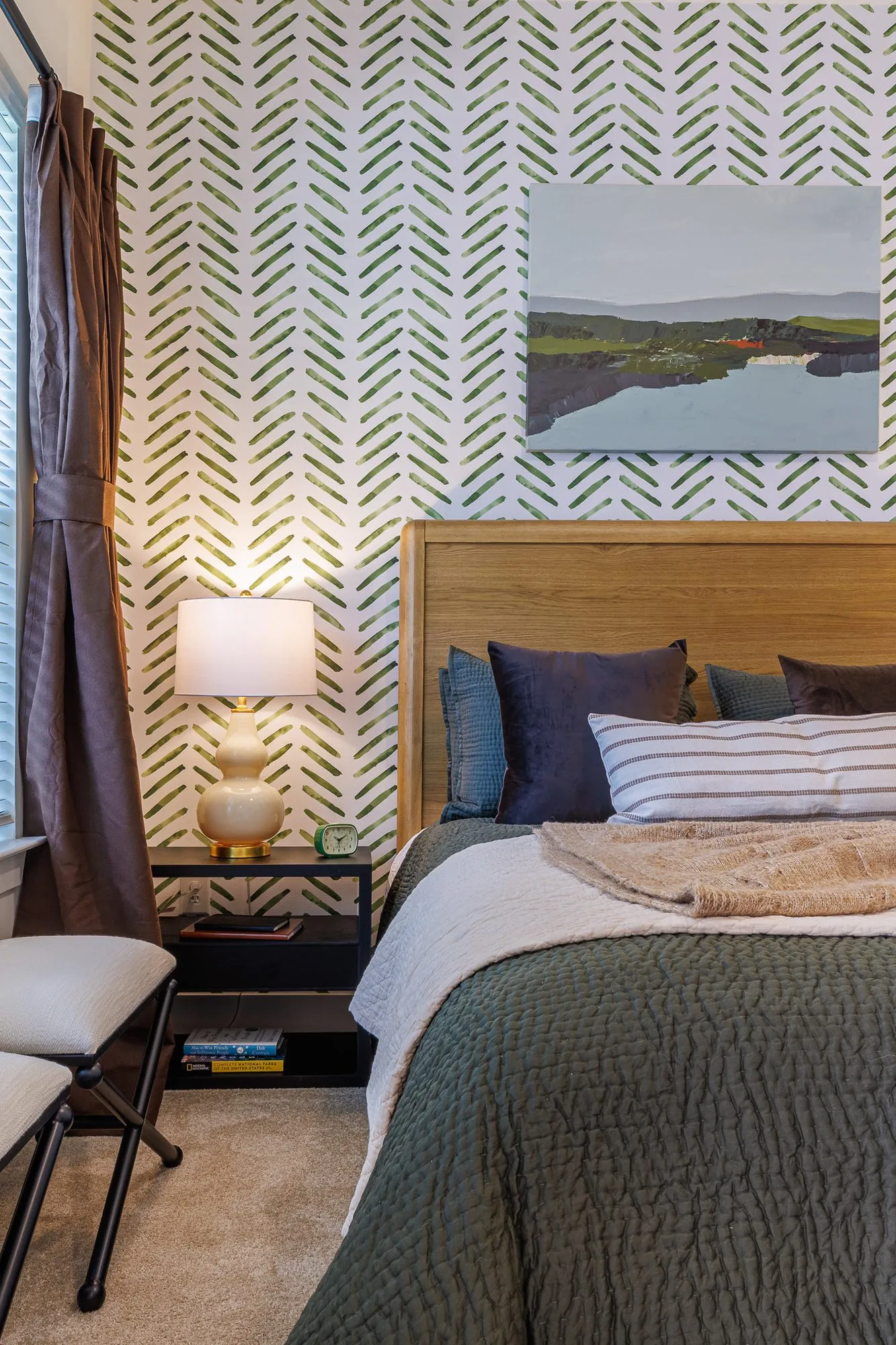 Multifamily model apartment bedroom with a wood headboard, green quilt and herringbone stencil wallpaper, landscape above