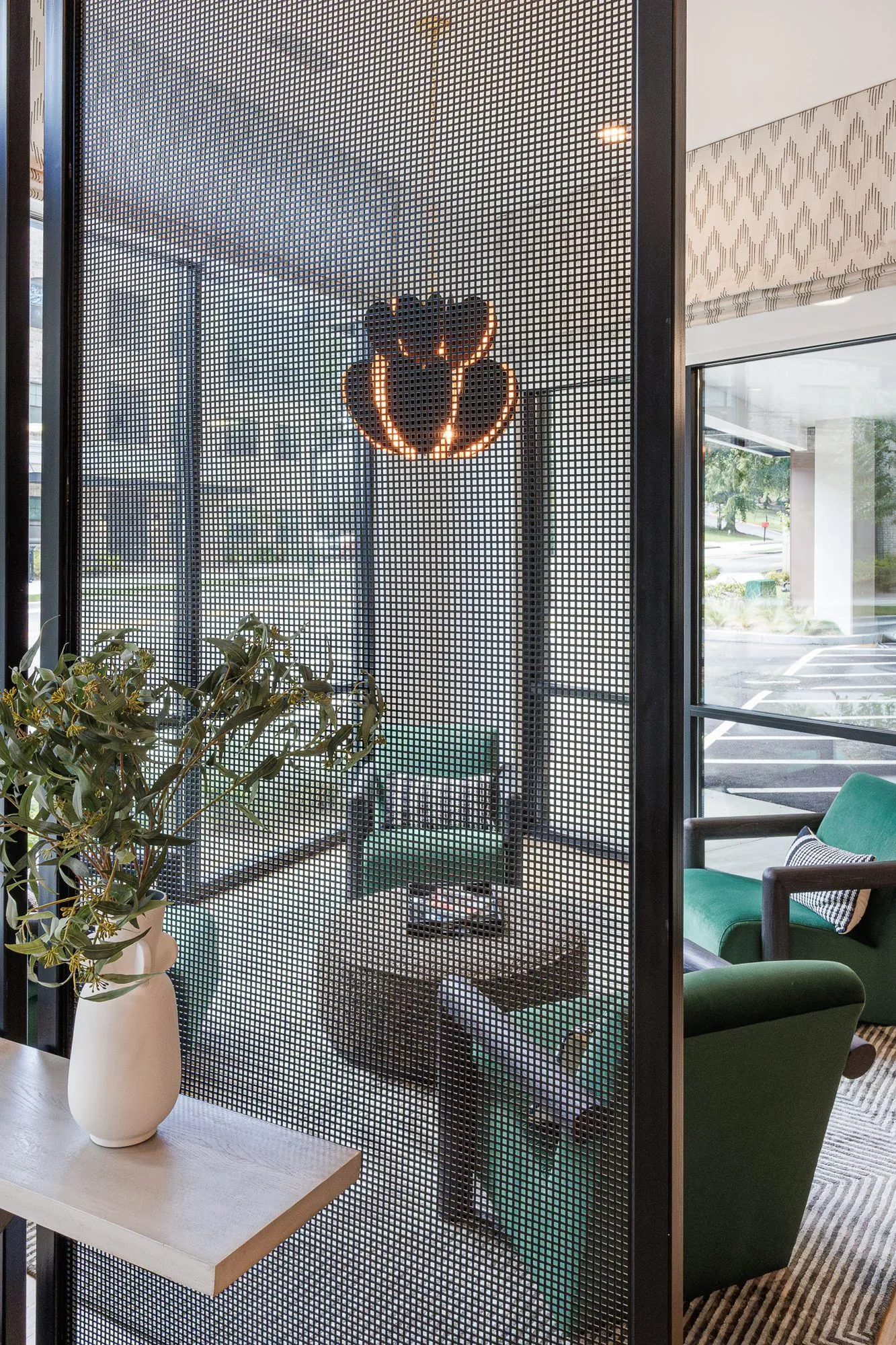 Multifamily amenity lounge with green velvet armchairs around a woven drum table, seen through a perforated metal screen