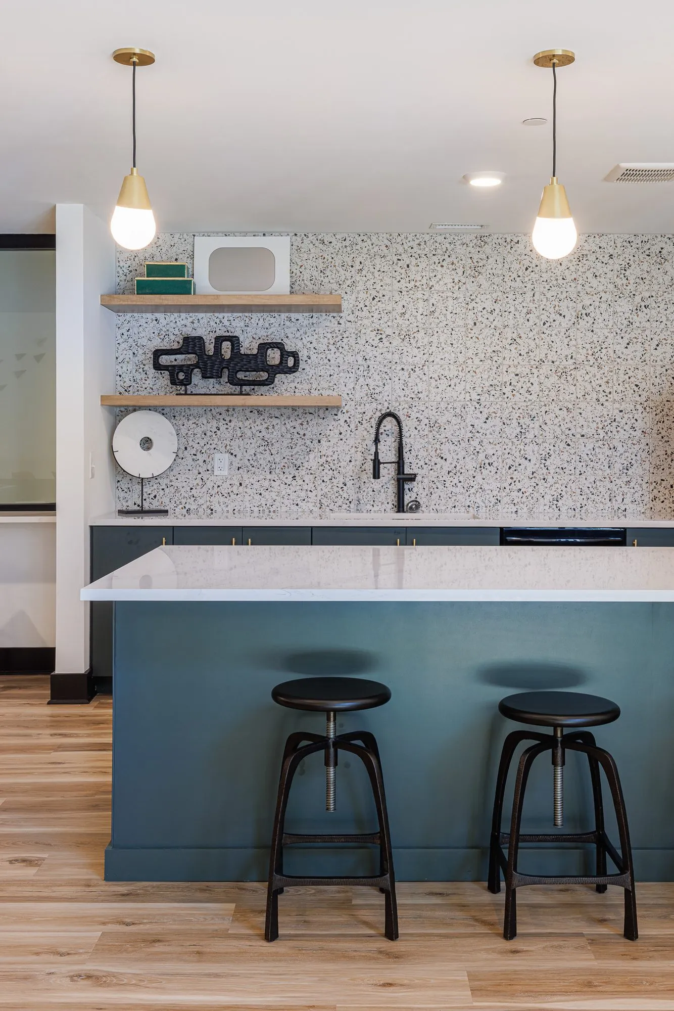 Multifamily kitchen island with a teal base and two black stools under brass pendants, terrazzo-look backsplash and shelves