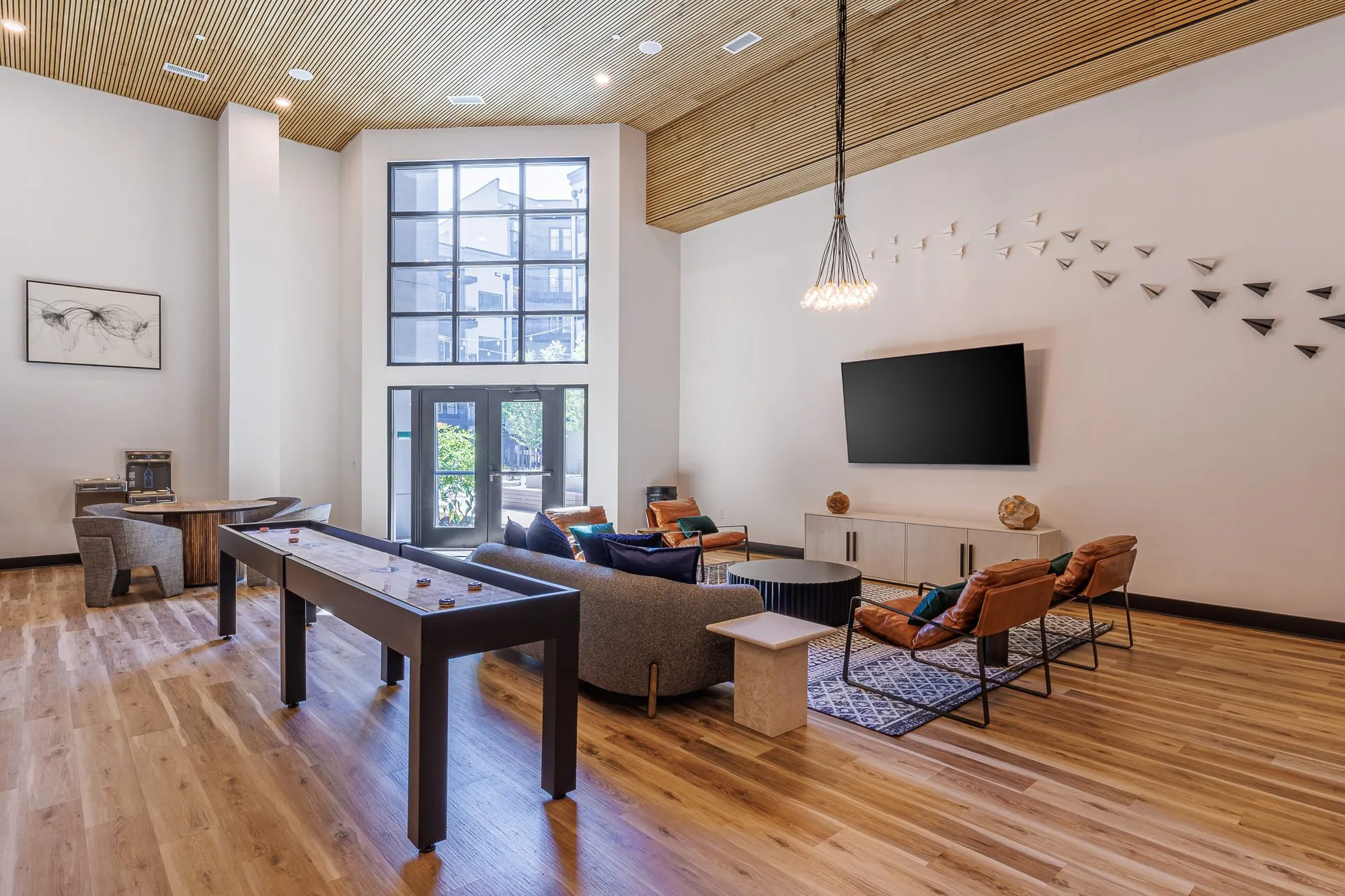 Multifamily amenity clubroom with a shuffleboard table, grey sofa and leather chairs facing a screen, paper planes above