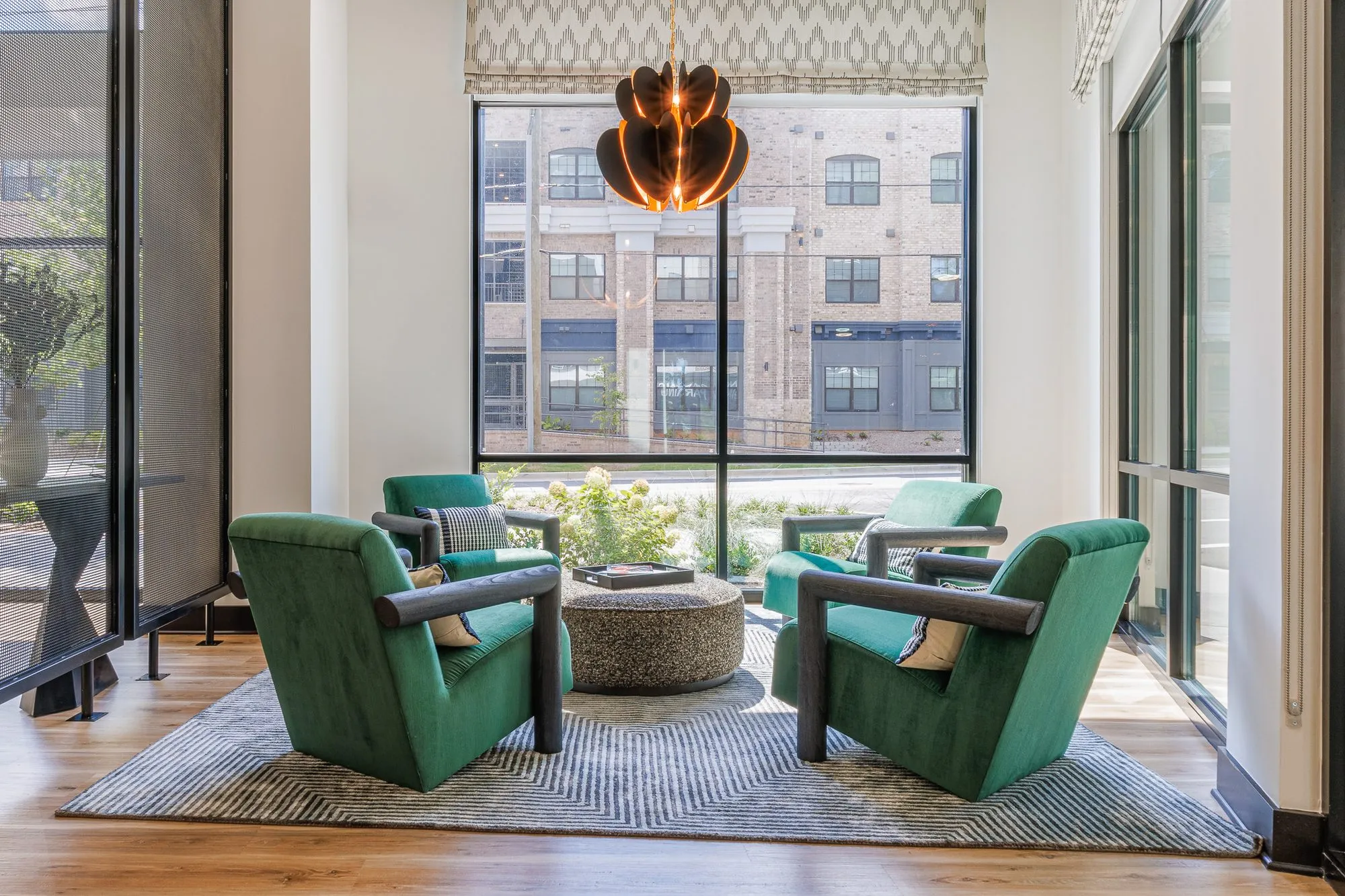 Multifamily interior detail: green velvet armchairs around a stone ottoman under a petal pendant, a tall window behind