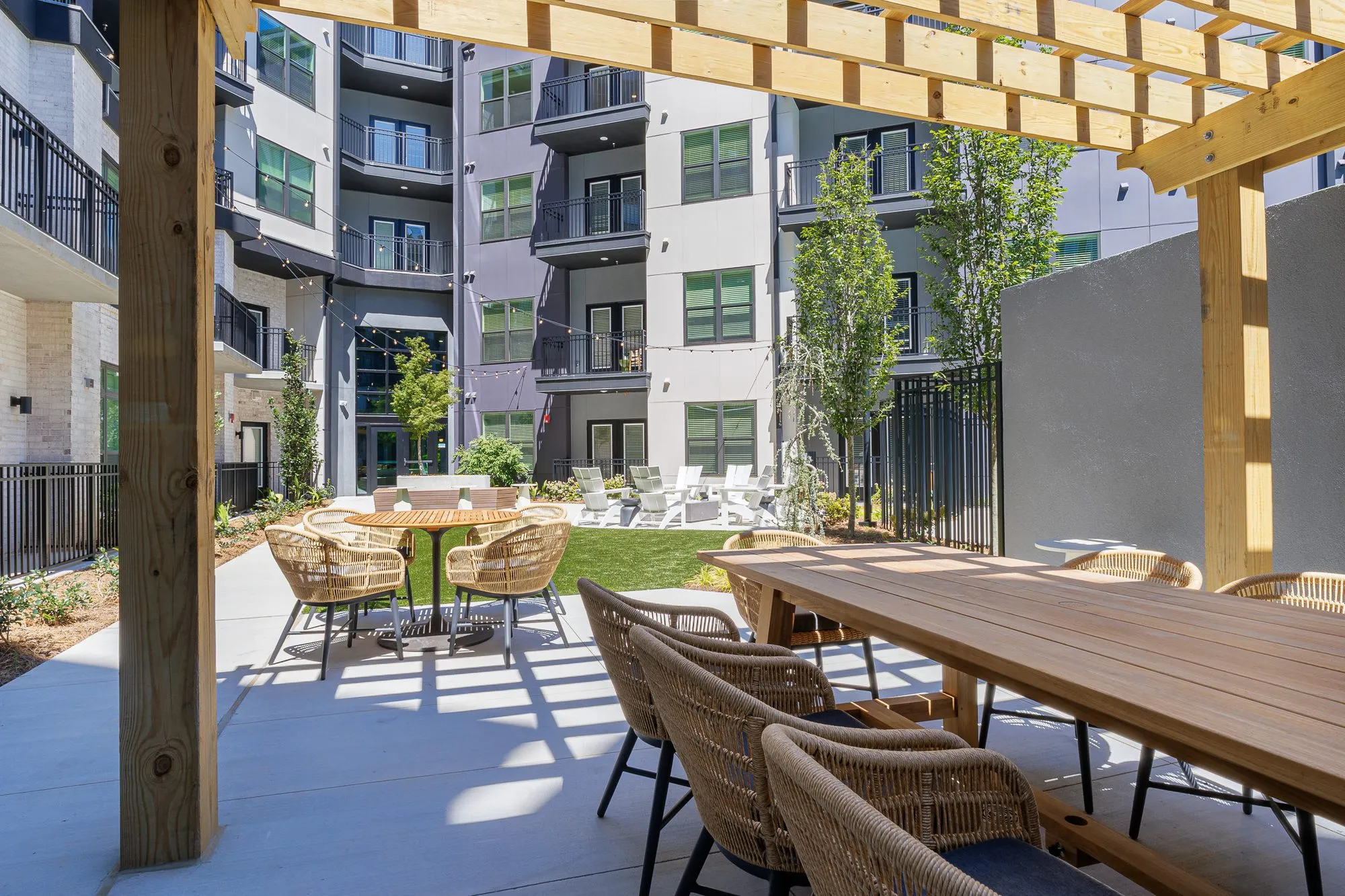 Multifamily amenity courtyard pergola over a long teak table with woven chairs, more dining sets and lawn beyond