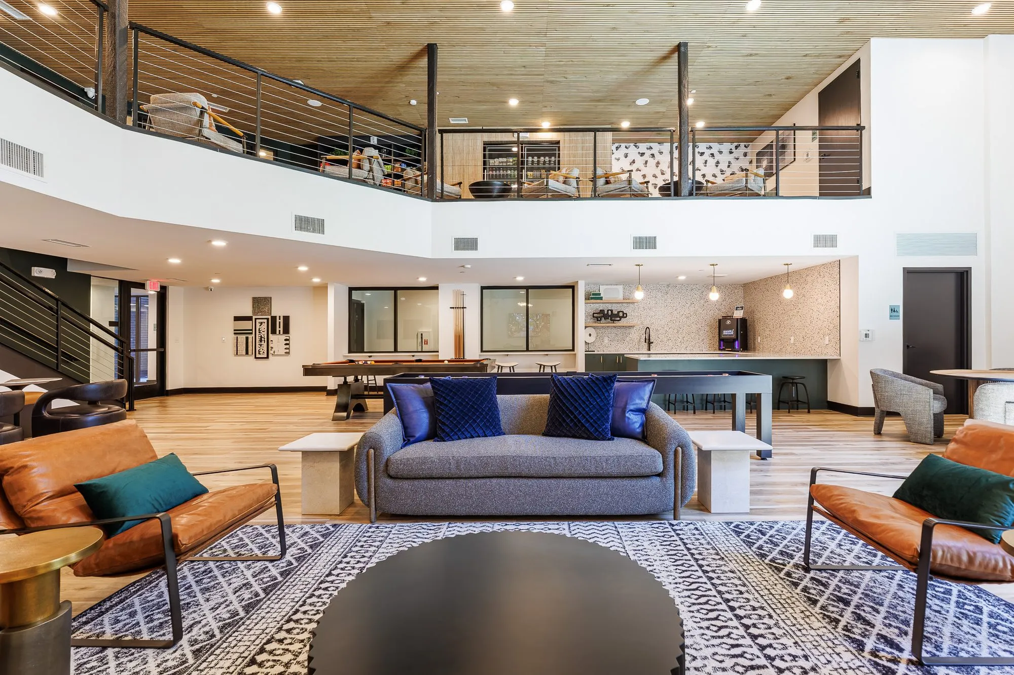 Multifamily amenity clubroom with cognac leather chairs and a grey sofa around a black drum table, mezzanine above
