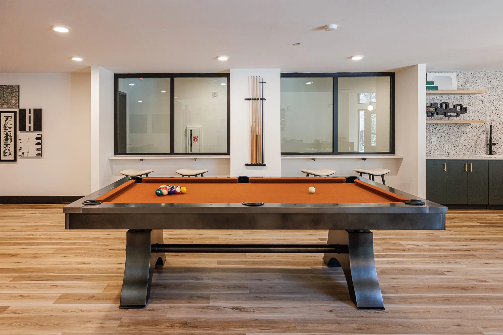 Multifamily amenity pool table with orange felt and a dark steel base before interior windows and a cue rack