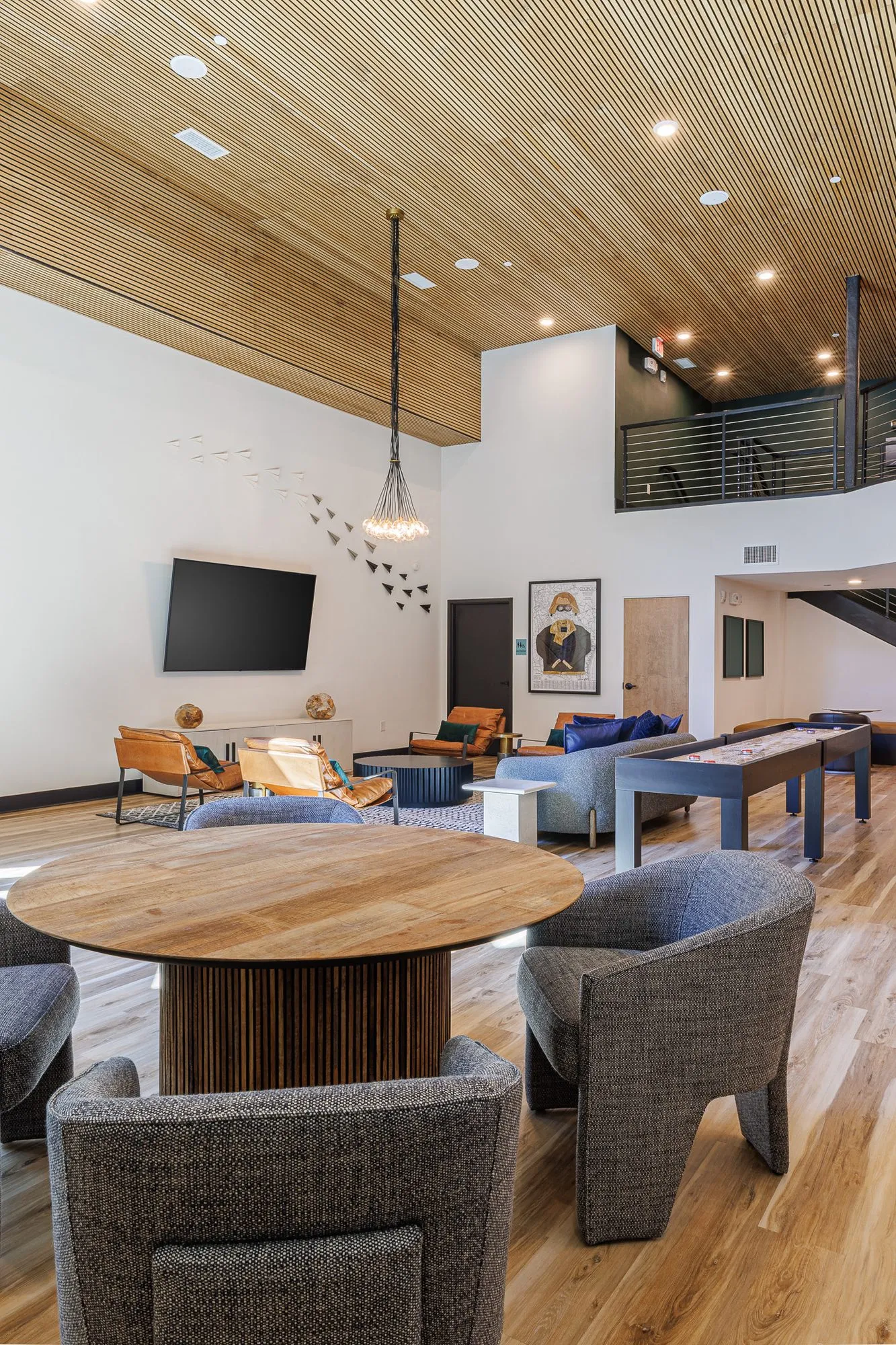 Multifamily amenity clubroom seen from a round wood table with grey chairs to the paper planes and mezzanine