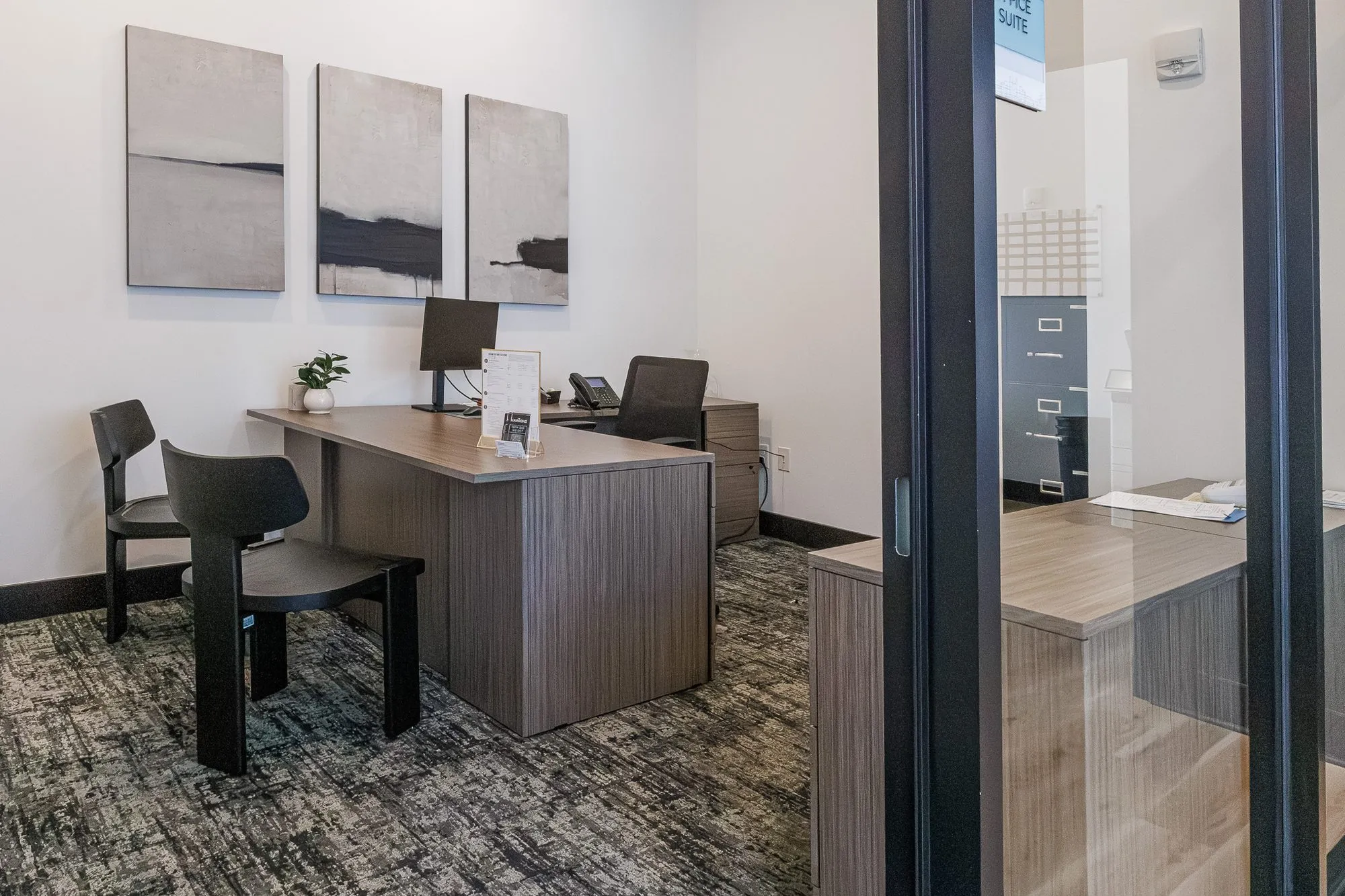 Multifamily amenity office with a wood desk and black chairs below three abstract paintings, seen through glass