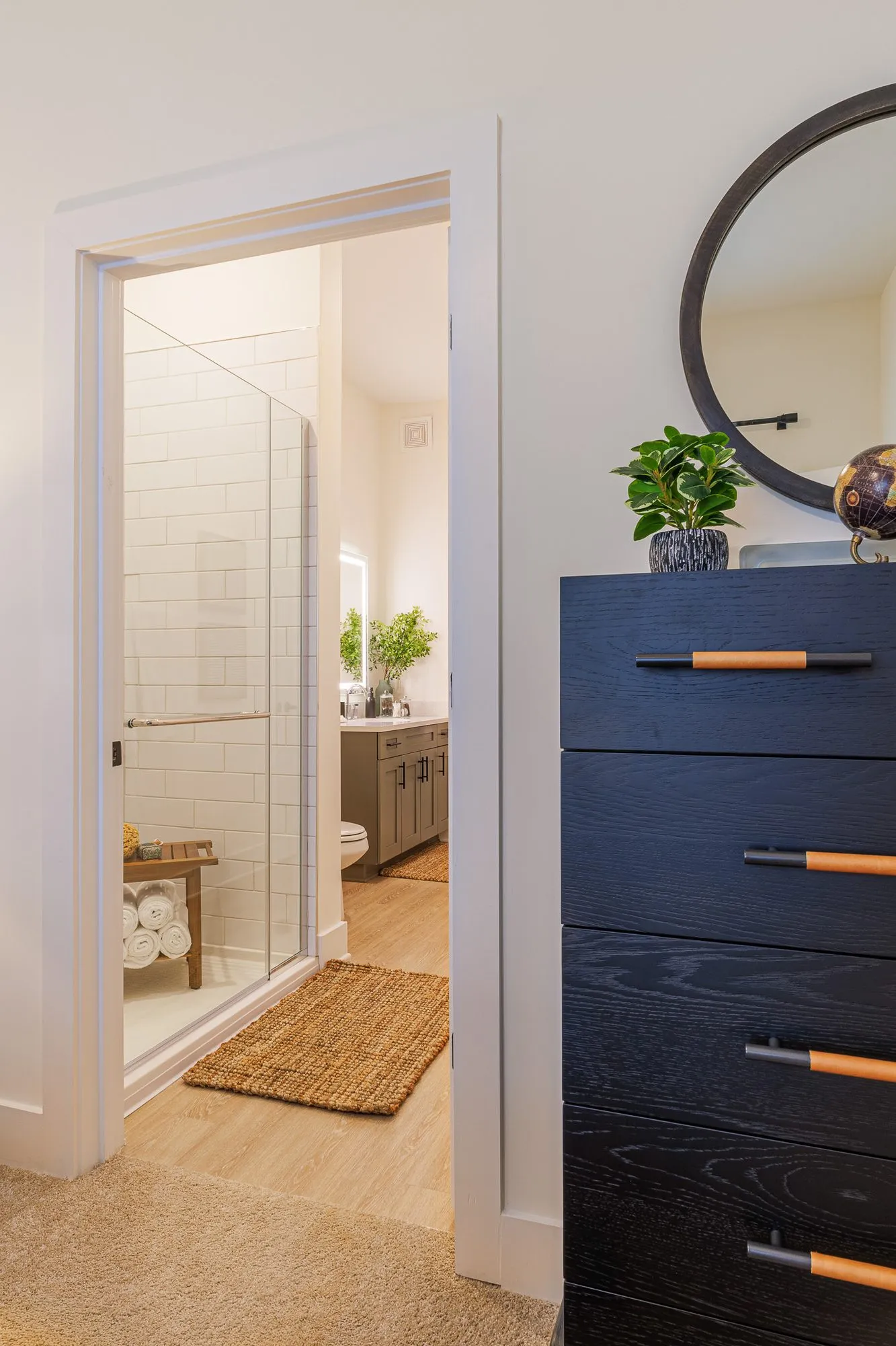 Multifamily model apartment bedroom with a black dresser and round mirror, the bathroom and glass shower beyond
