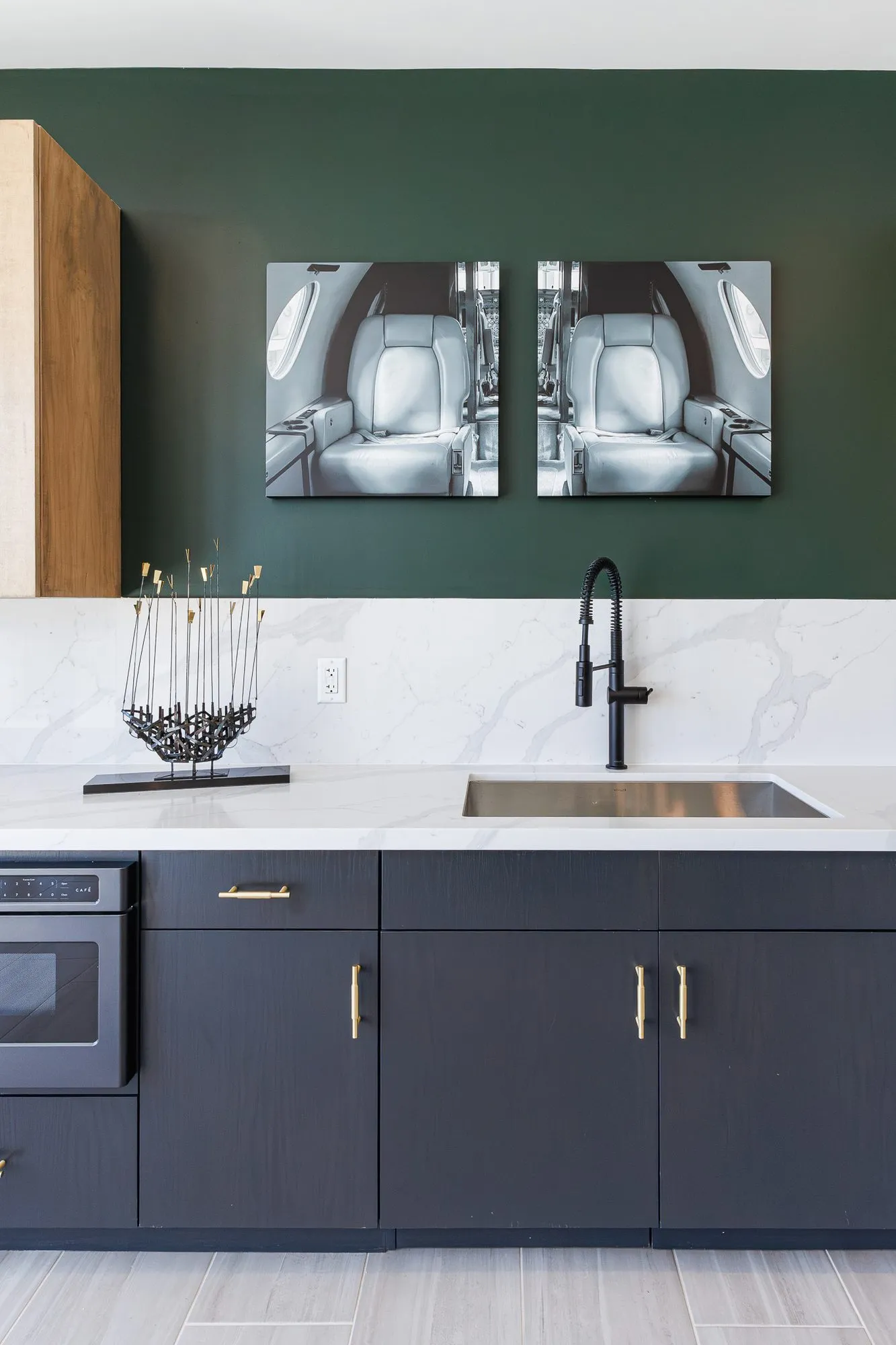 Multifamily amenity kitchenette with charcoal cabinets and a black faucet below two aircraft cabin photographs on green