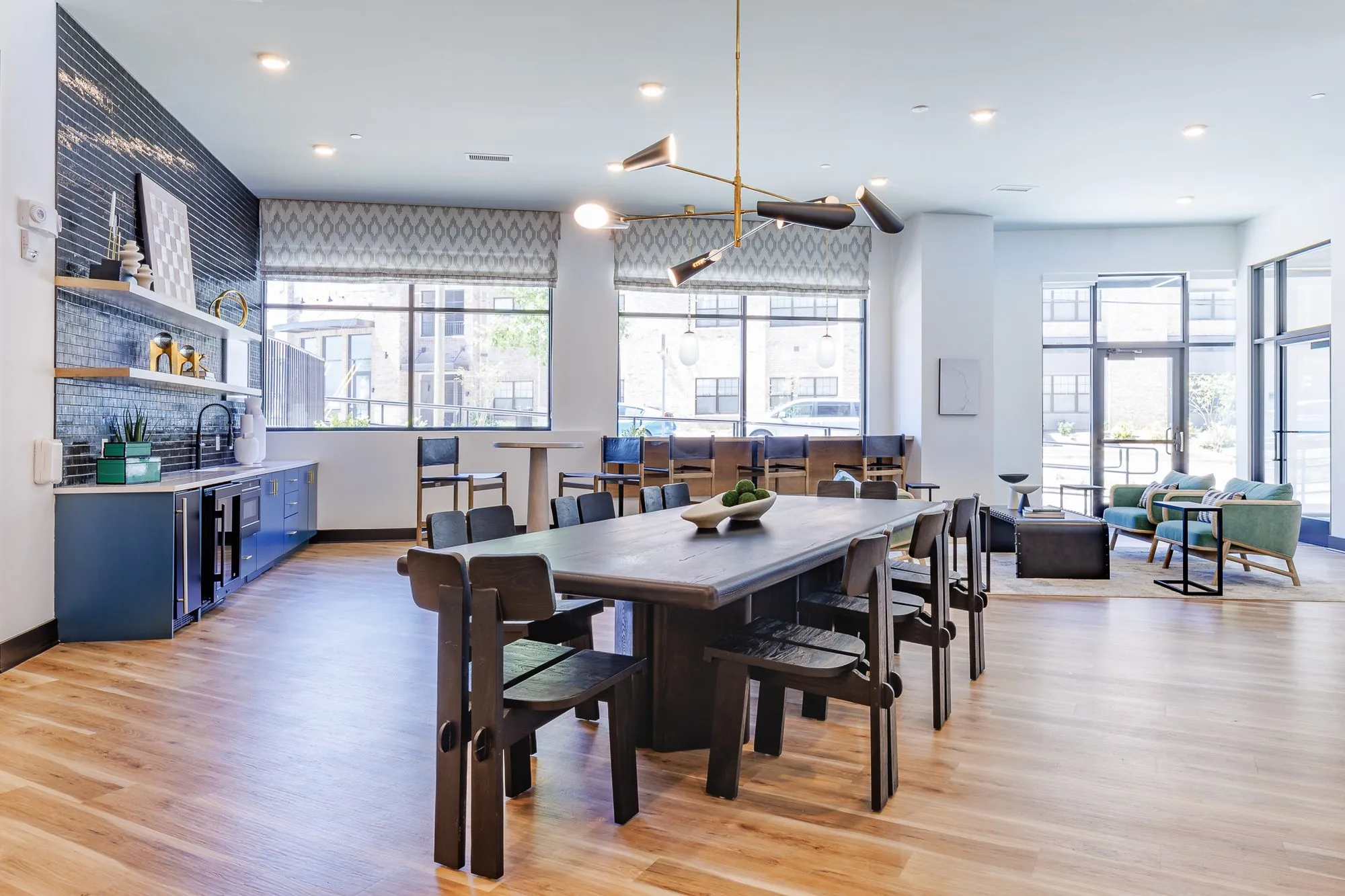 Multifamily amenity clubroom with a long dark table under a brass stick chandelier, blue cabinetry and black tile wall