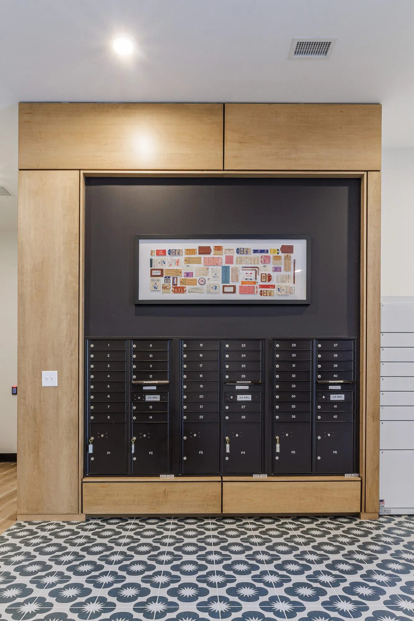 Multifamily amenity mailboxes set in an oak surround below a framed collage, patterned cement tile floor