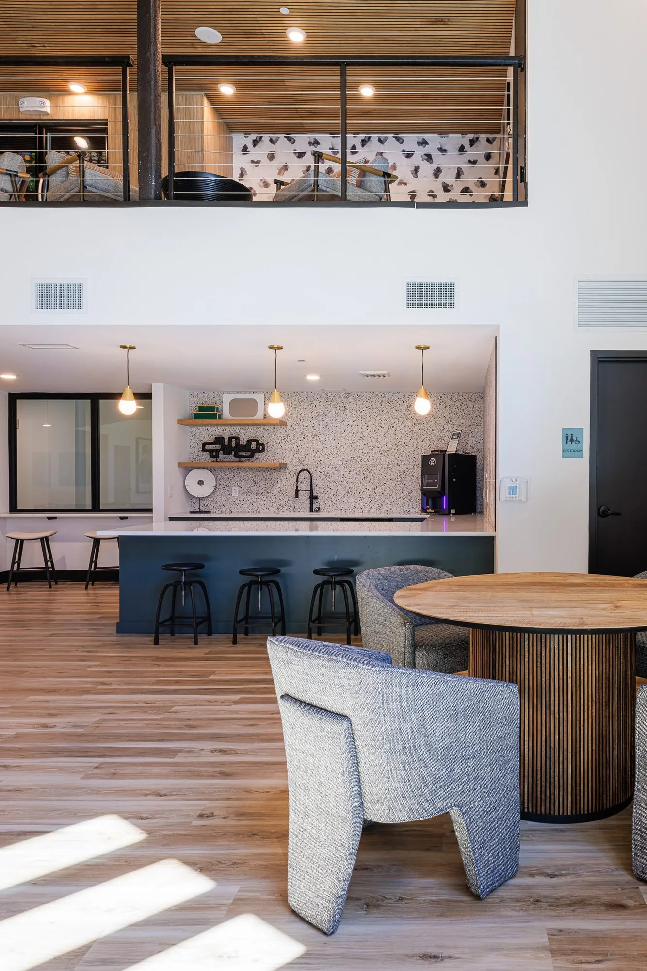 Multifamily kitchen with a teal island and black stools beneath the mezzanine, grey chairs at a fluted table in front