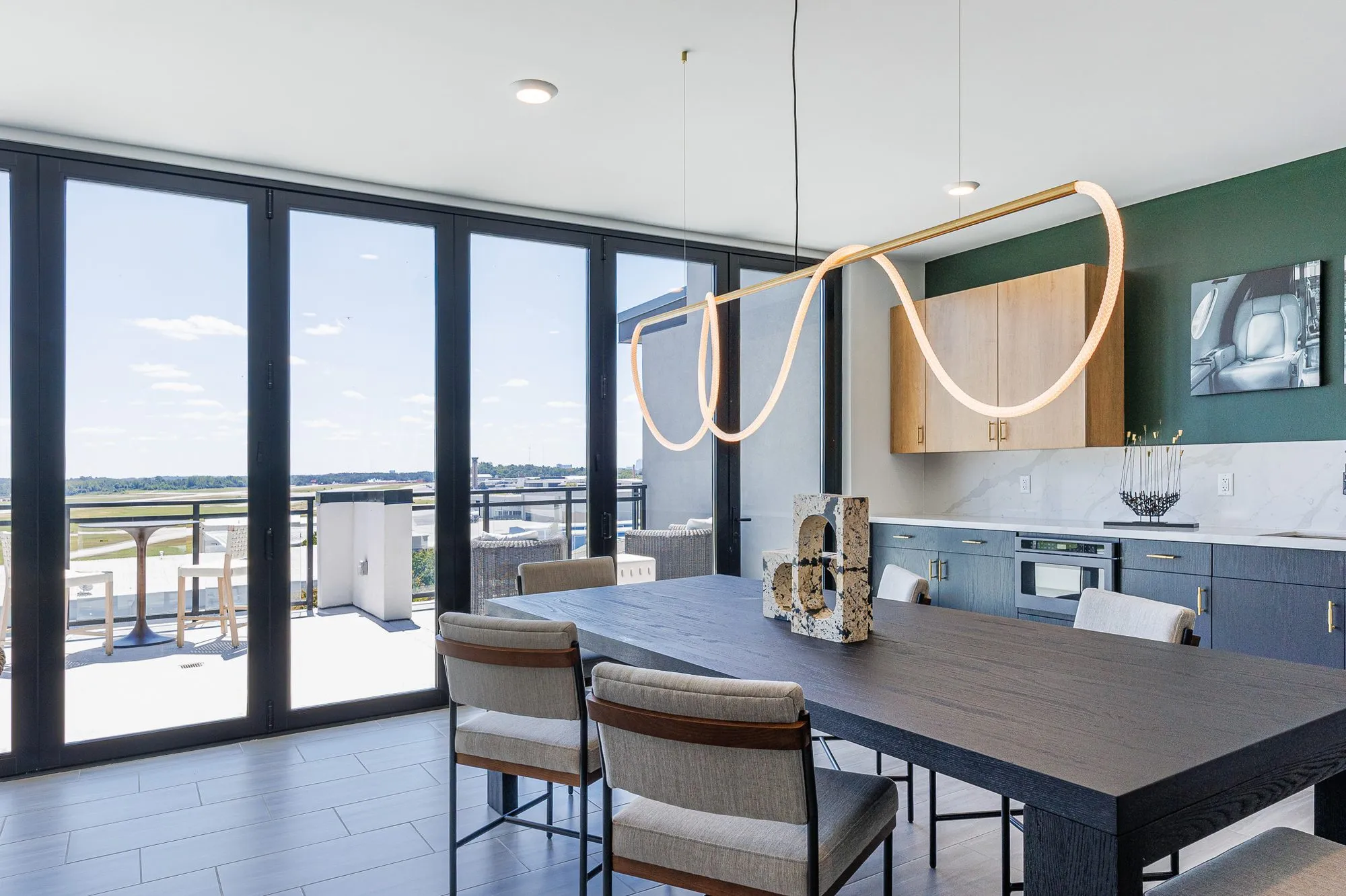 Multifamily amenity clubroom, a long view down the dark dining table under the rope light, folding glass walls to the terrace