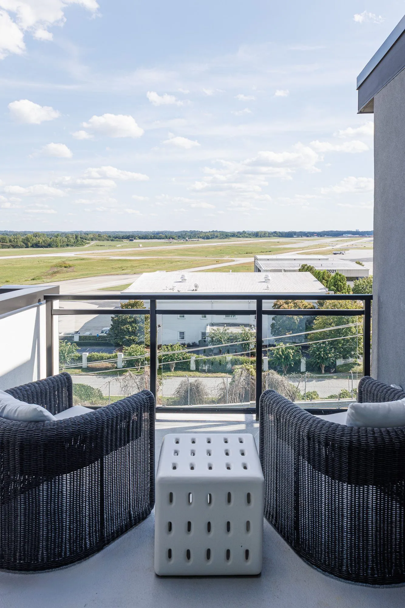 Multifamily apartment balcony with wicker lounge chairs and a perforated cube table, the airfield and sky beyond