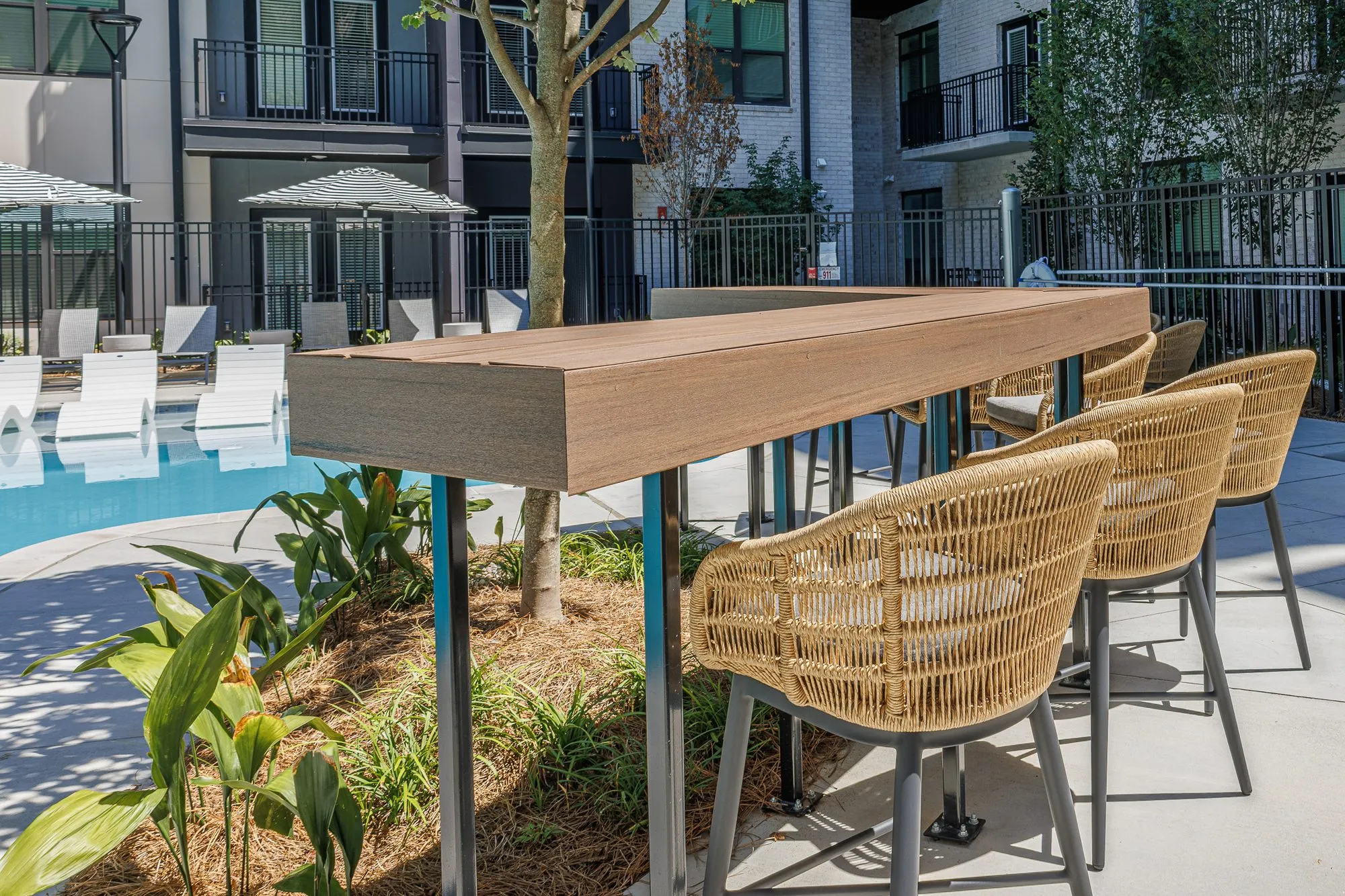 Multifamily amenity pool detail: woven counter chairs at a high wood table set in a planted bed beside the pool