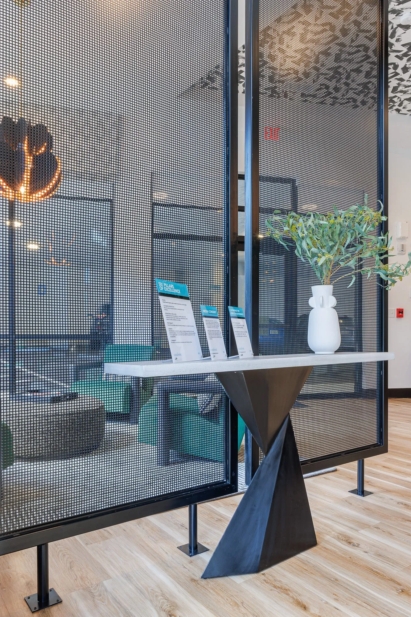 Multifamily interior detail: black twisted pedestal console with brochures beside a perforated metal screen, green armchairs