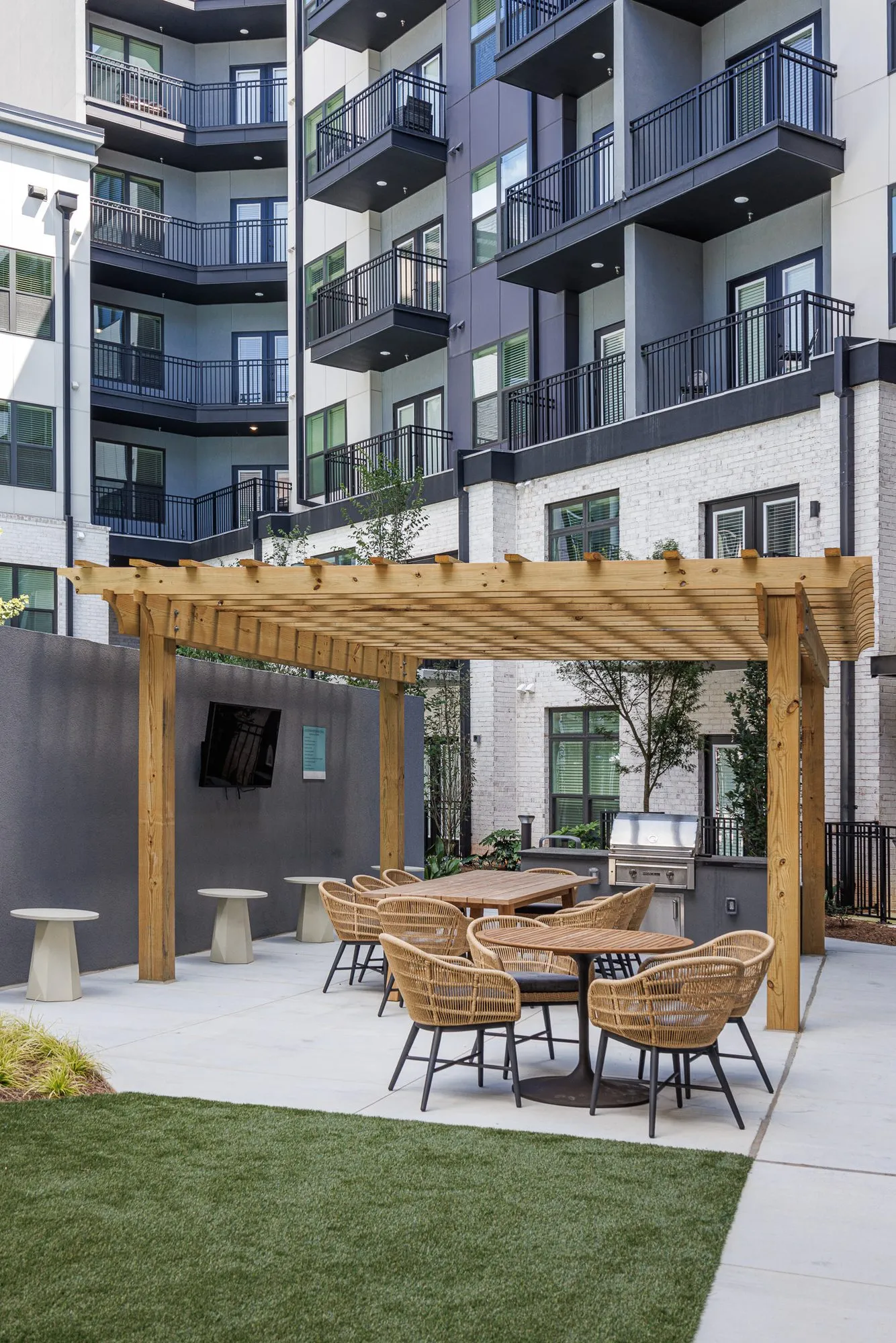 Multifamily amenity courtyard pergola with woven dining chairs, an outdoor grill and wall screen below residential balconies