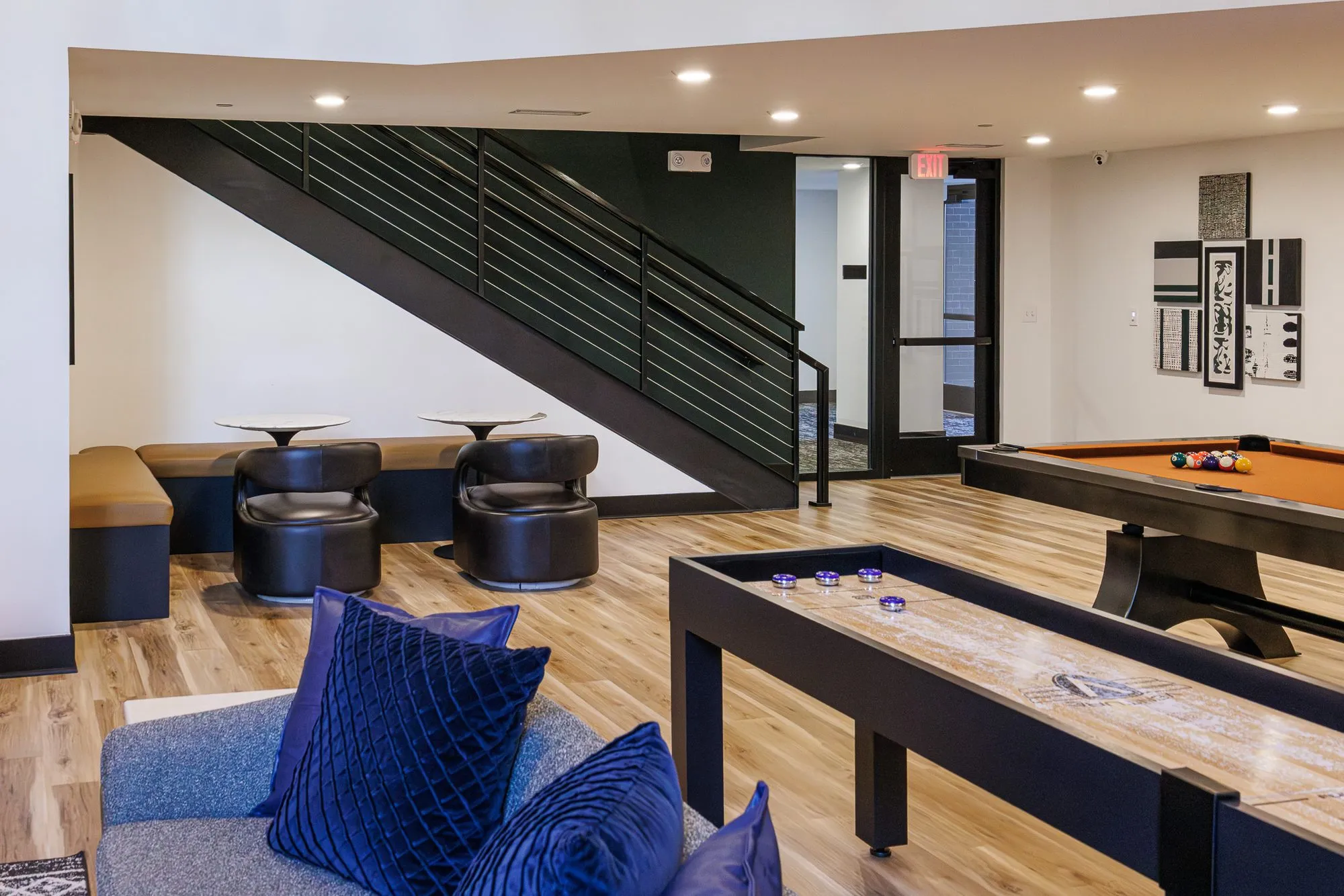 Multifamily amenity game area with a shuffleboard table, pool table and leather tub chairs at a banquette under the stair