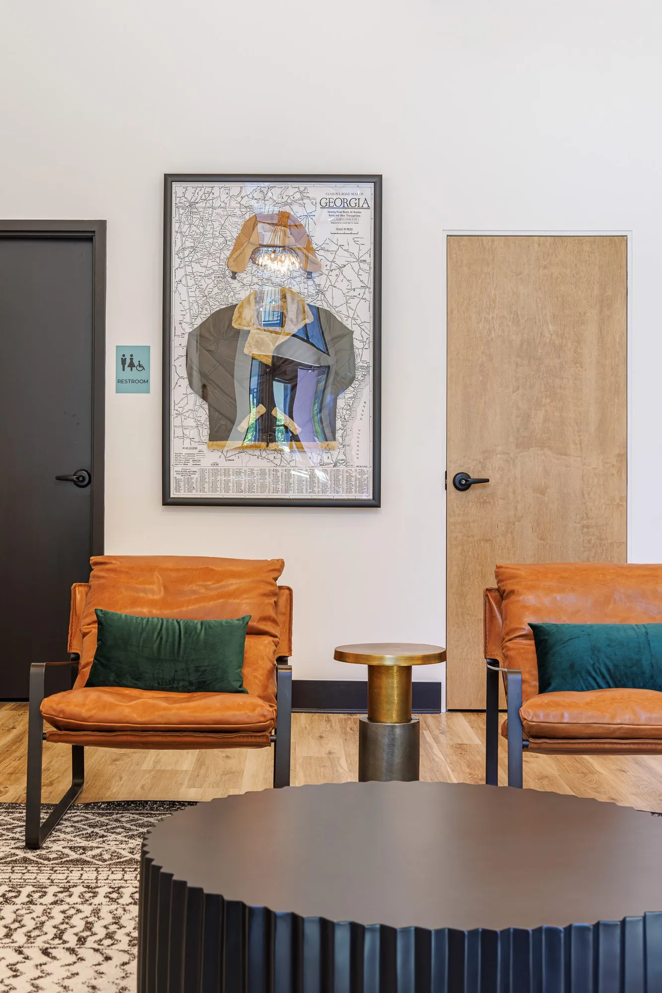 Multifamily interior detail: cognac leather chairs with green pillows and a brass side table below a framed aviator collage