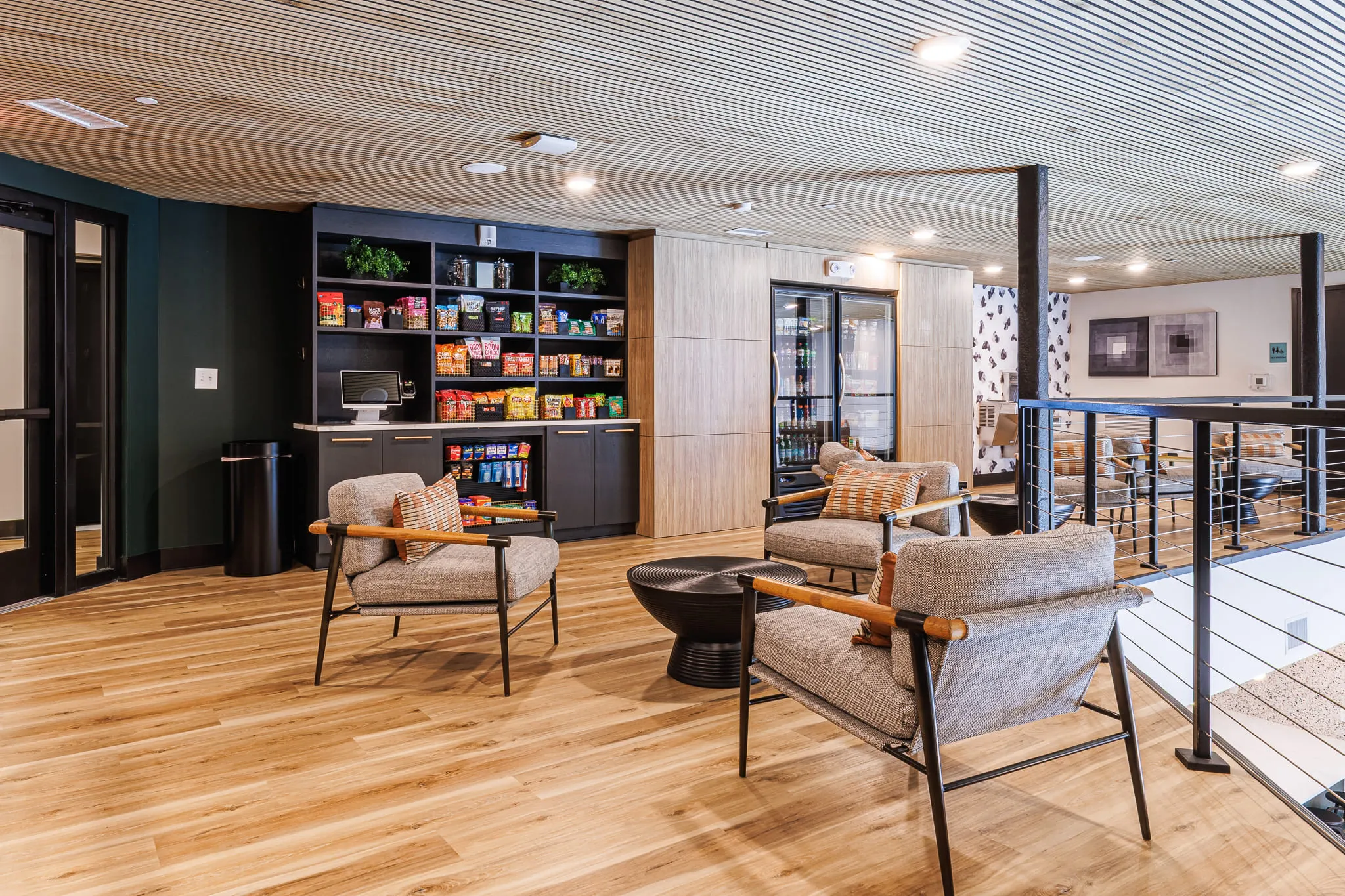 Multifamily amenity snack market with stocked dark shelving and a drinks fridge, grey armchairs with wood arms in front