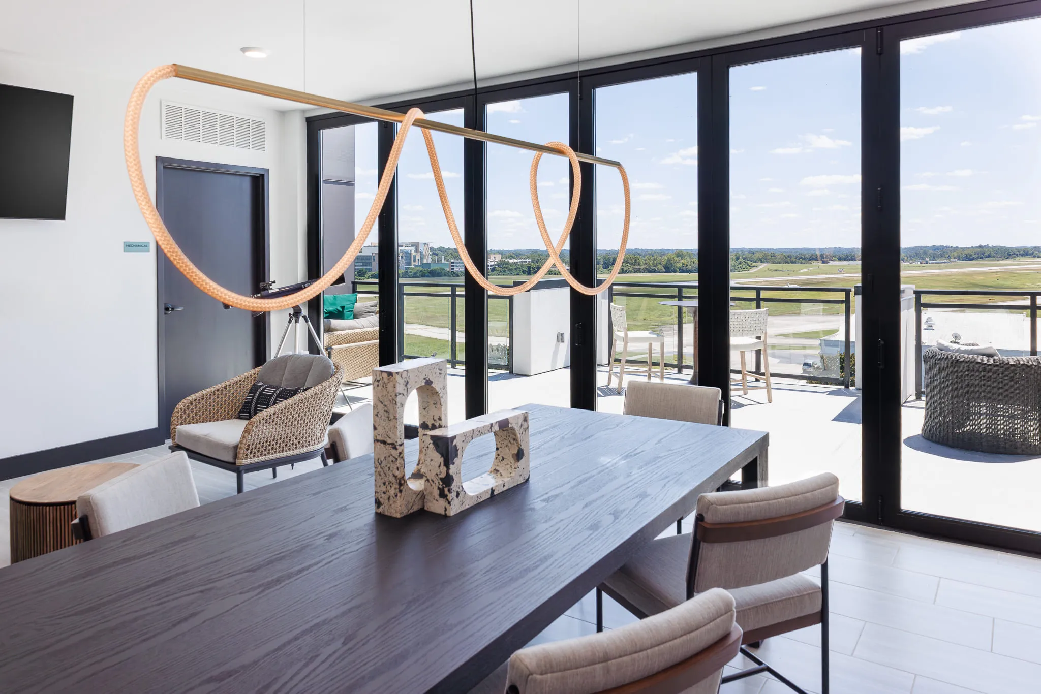 Multifamily amenity communal dining table under a looping rope light, the airfield beyond the glass