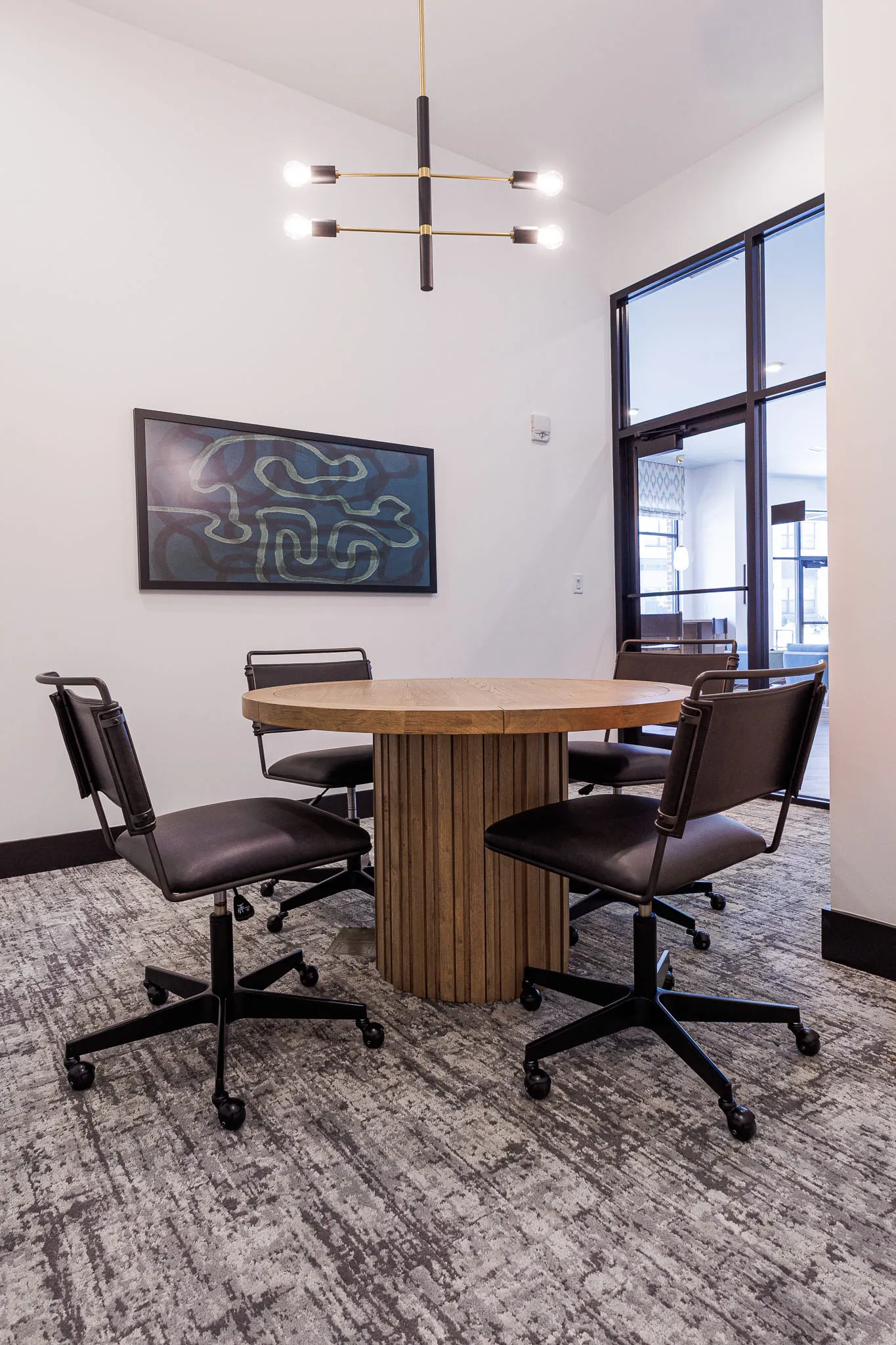 Multifamily amenity conference room with a round fluted wood table and black leather task chairs below a framed line artwork