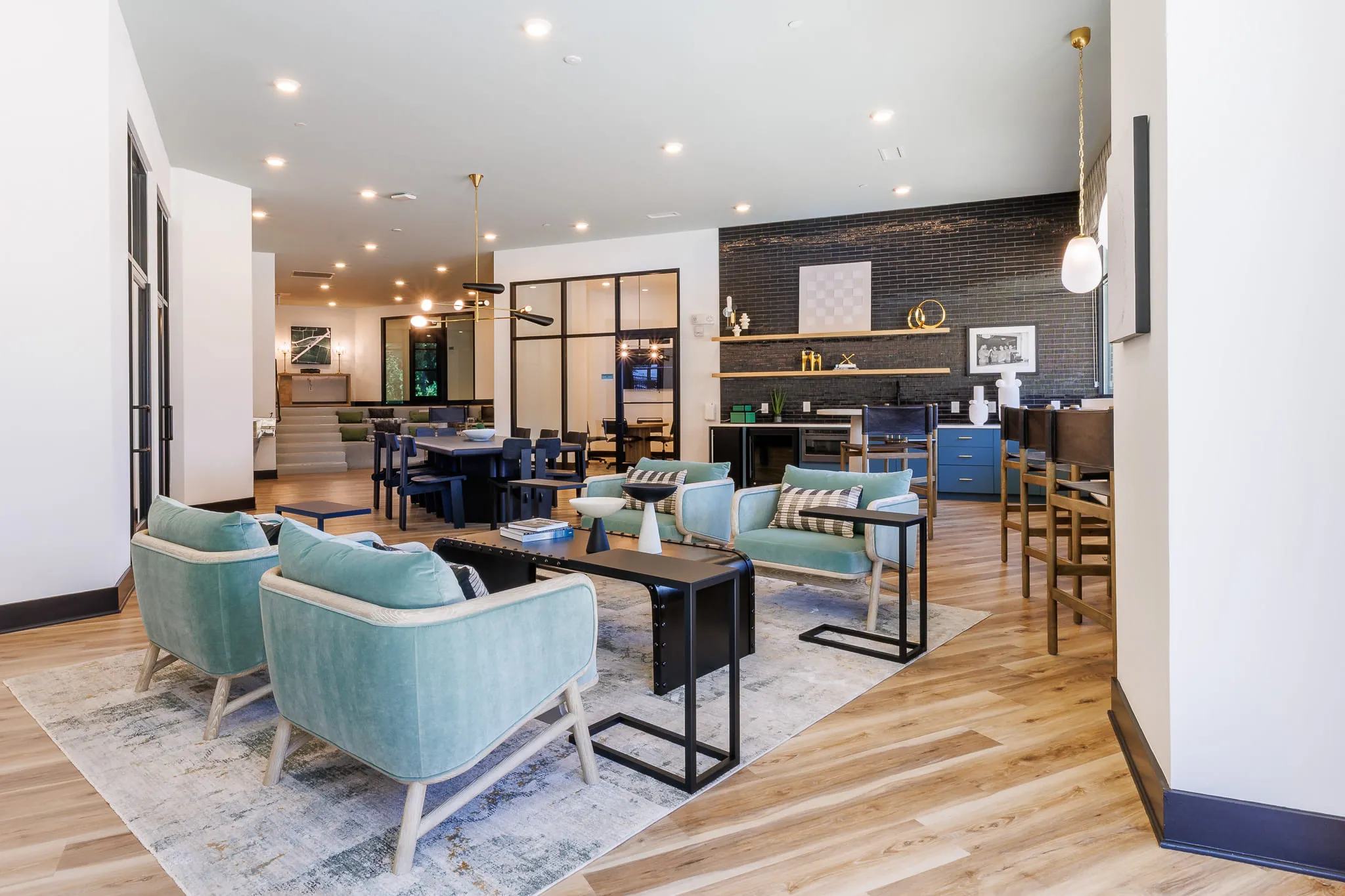Multifamily amenity lounge with teal armchairs and C-tables before a glass-walled room, dining tables and the coffee bar
