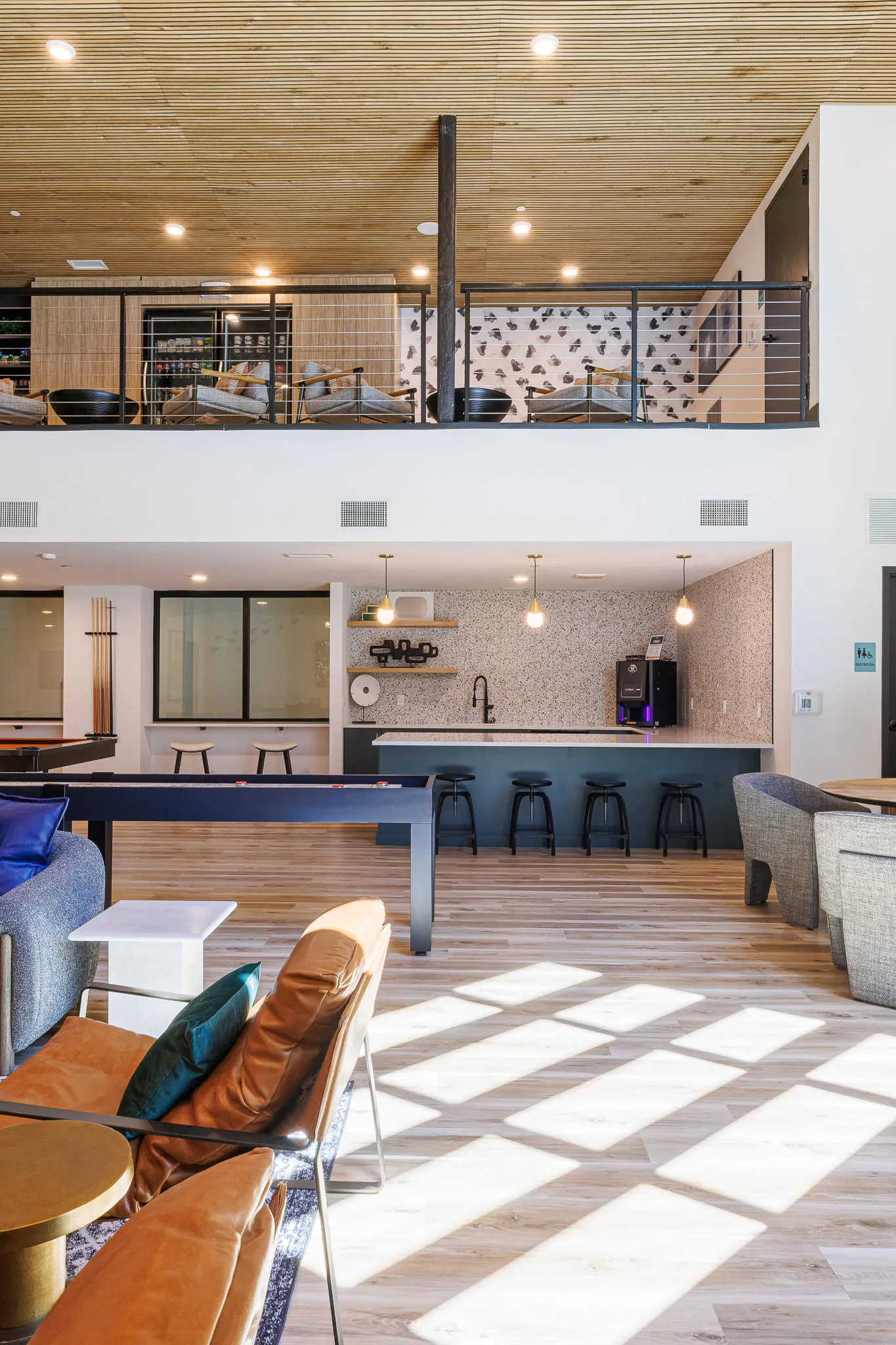 Multifamily amenity game room with a shuffleboard and pool table, a navy counter with black stools below the mezzanine