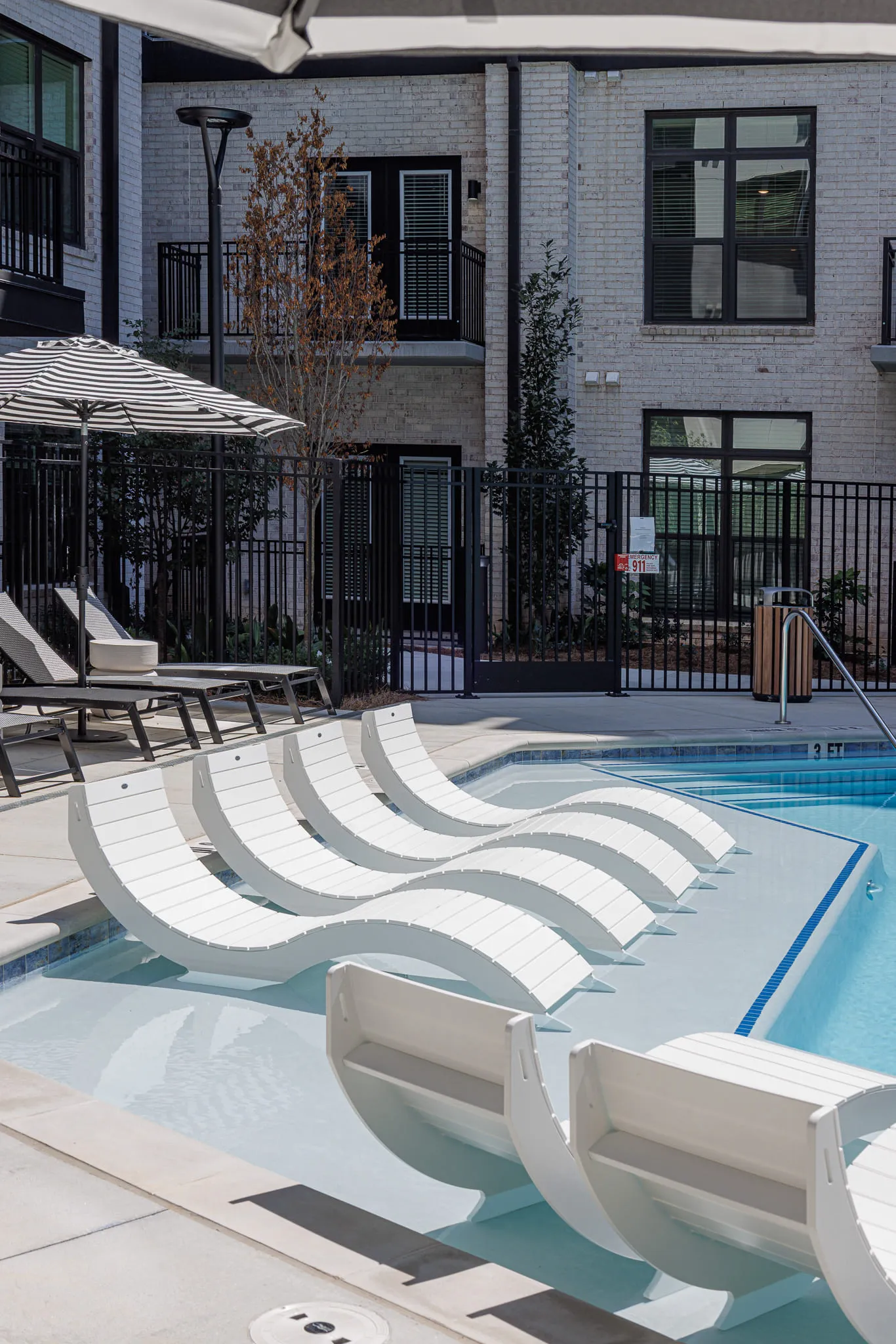 Multifamily amenity pool with white curved chaises in the shallow ledge beside a striped umbrella