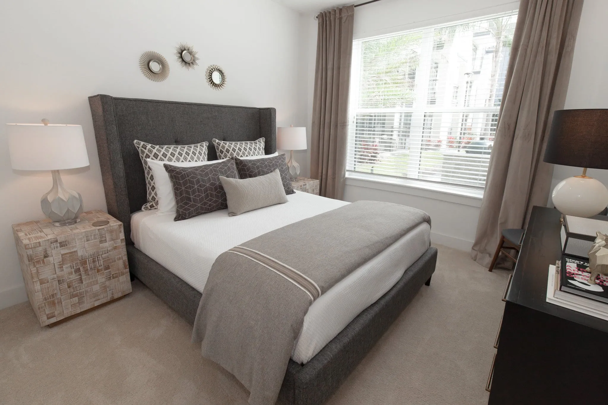 Multifamily apartment model bedroom with a grey upholstered headboard and layered bedding by a whitewashed block nightstand