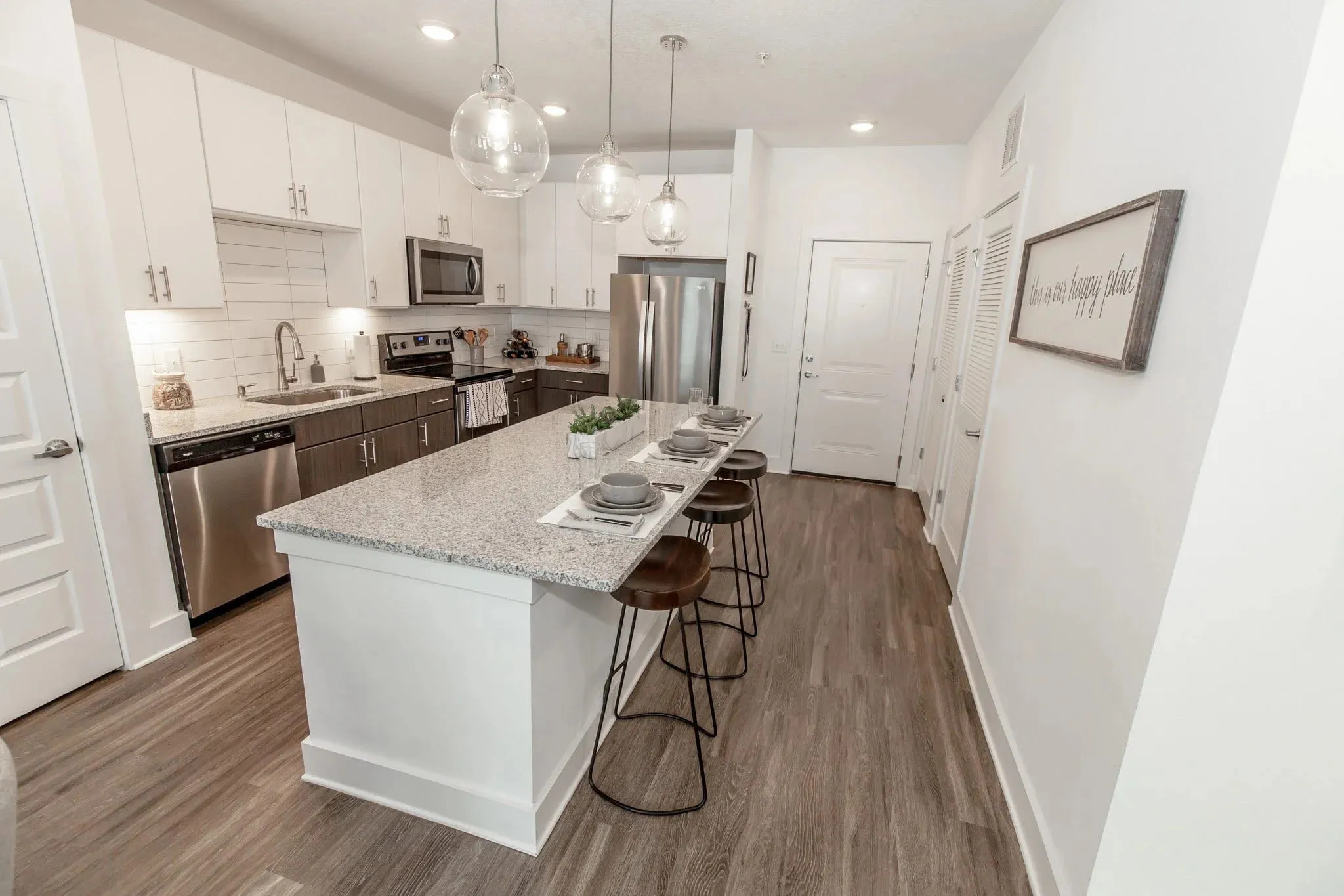 Multifamily apartment model kitchen with white cabinetry and a granite island, wood-seat stools under glass globe pendants