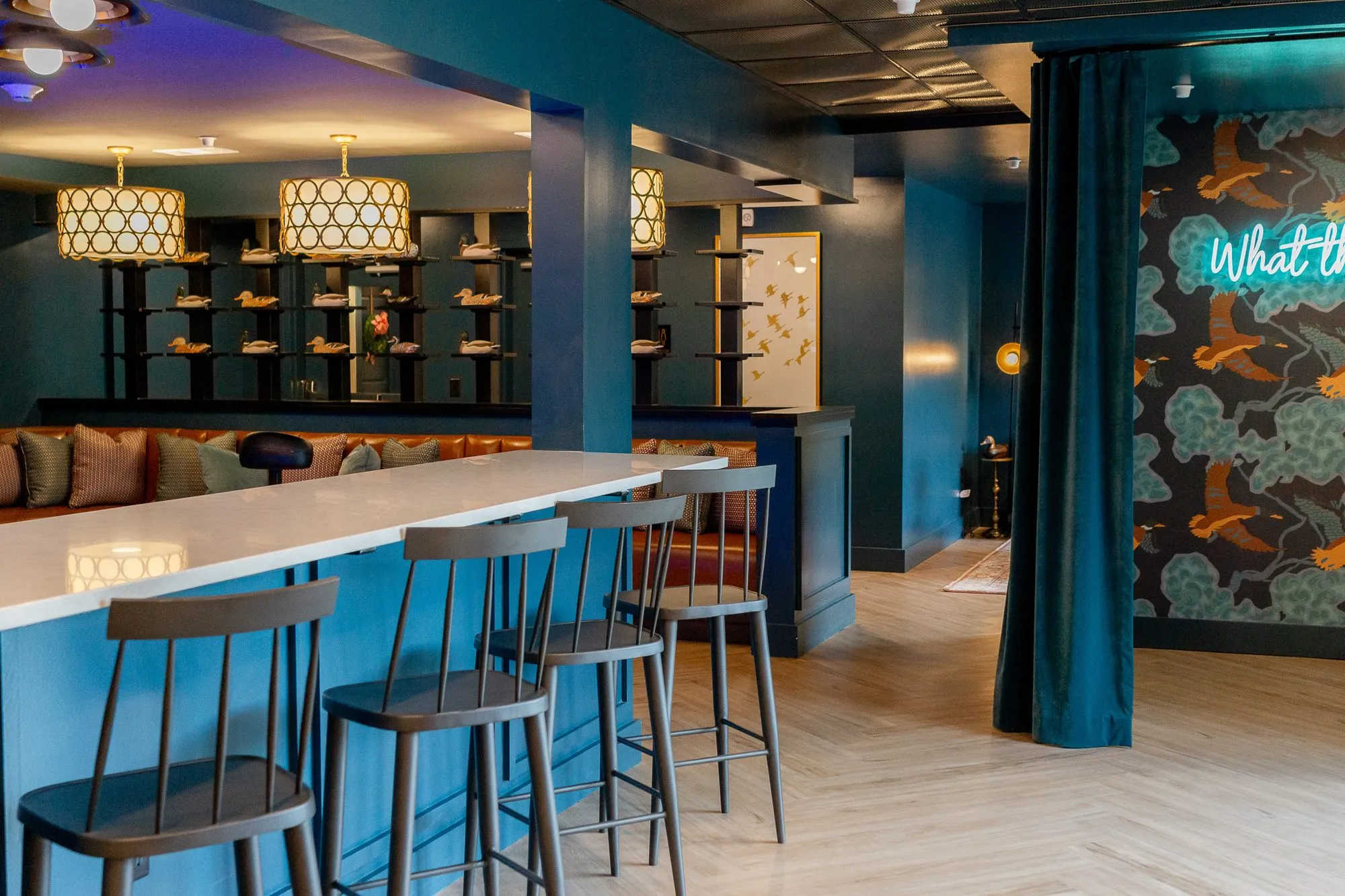 Multifamily amenity bar with wood stools at a white counter under ring-pattern drum pendants, a teal curtain and neon beyond