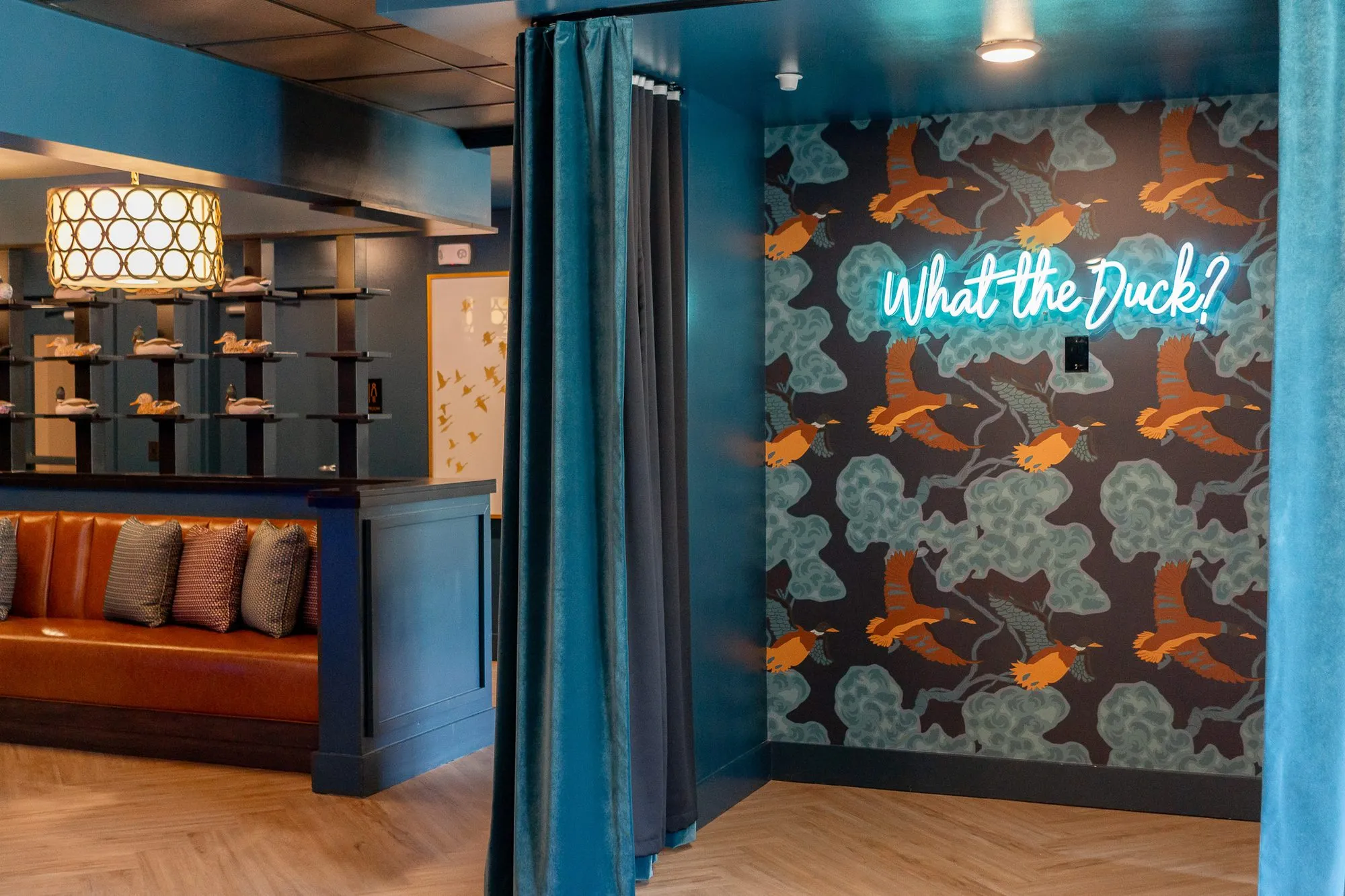 Multifamily amenity alcove with teal velvet curtains framing a neon What the Duck? sign on dark duck-print wallpaper