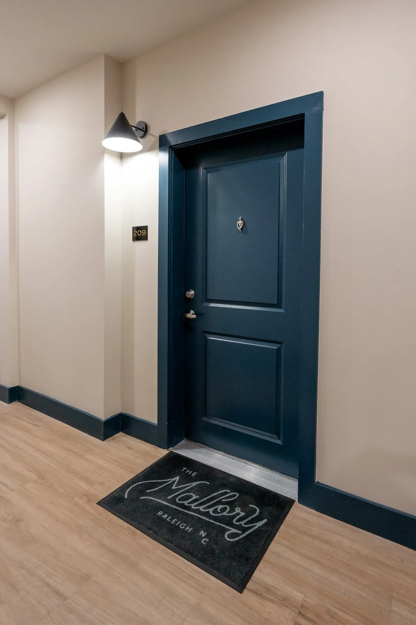 Multifamily residential corridor with a navy apartment door, a black sconce and a doormat bearing a script logo