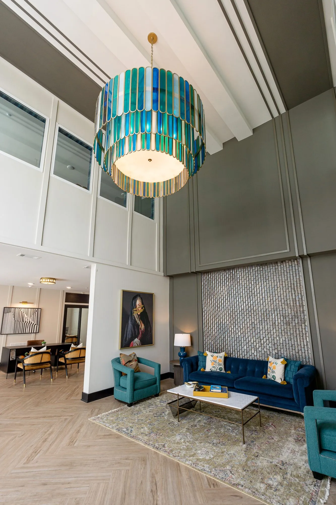 Multifamily amenity lobby with the glass chandelier above a navy sofa and teal chairs, the leasing area through the opening