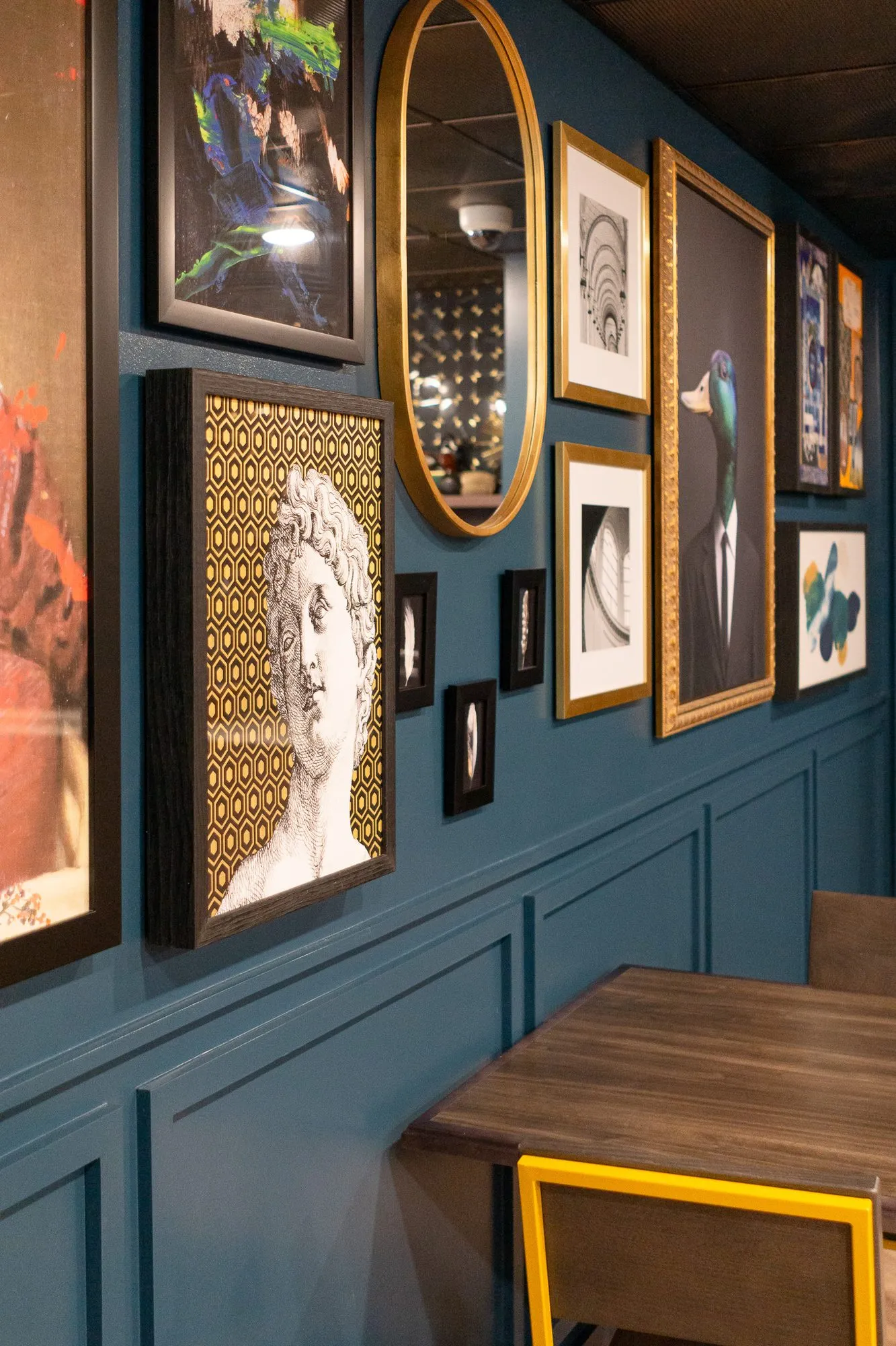 Multifamily amenity gallery wall on dark teal panelling with a bust print, an oval mirror and photographs above a wood table
