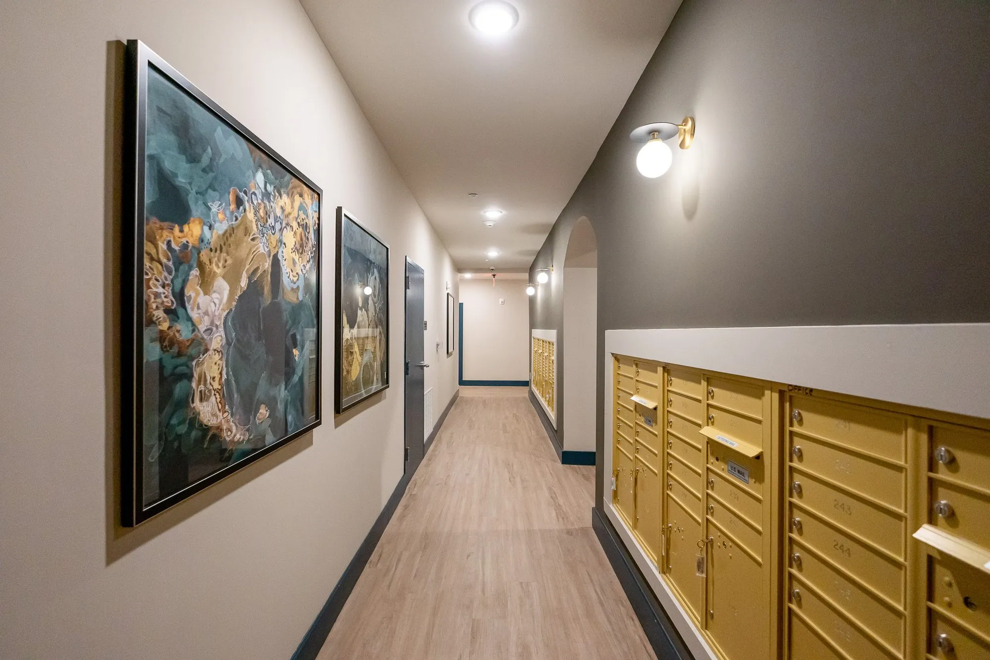 Multifamily amenity mail corridor with brass mailboxes and large framed abstract paintings, an arched opening beyond