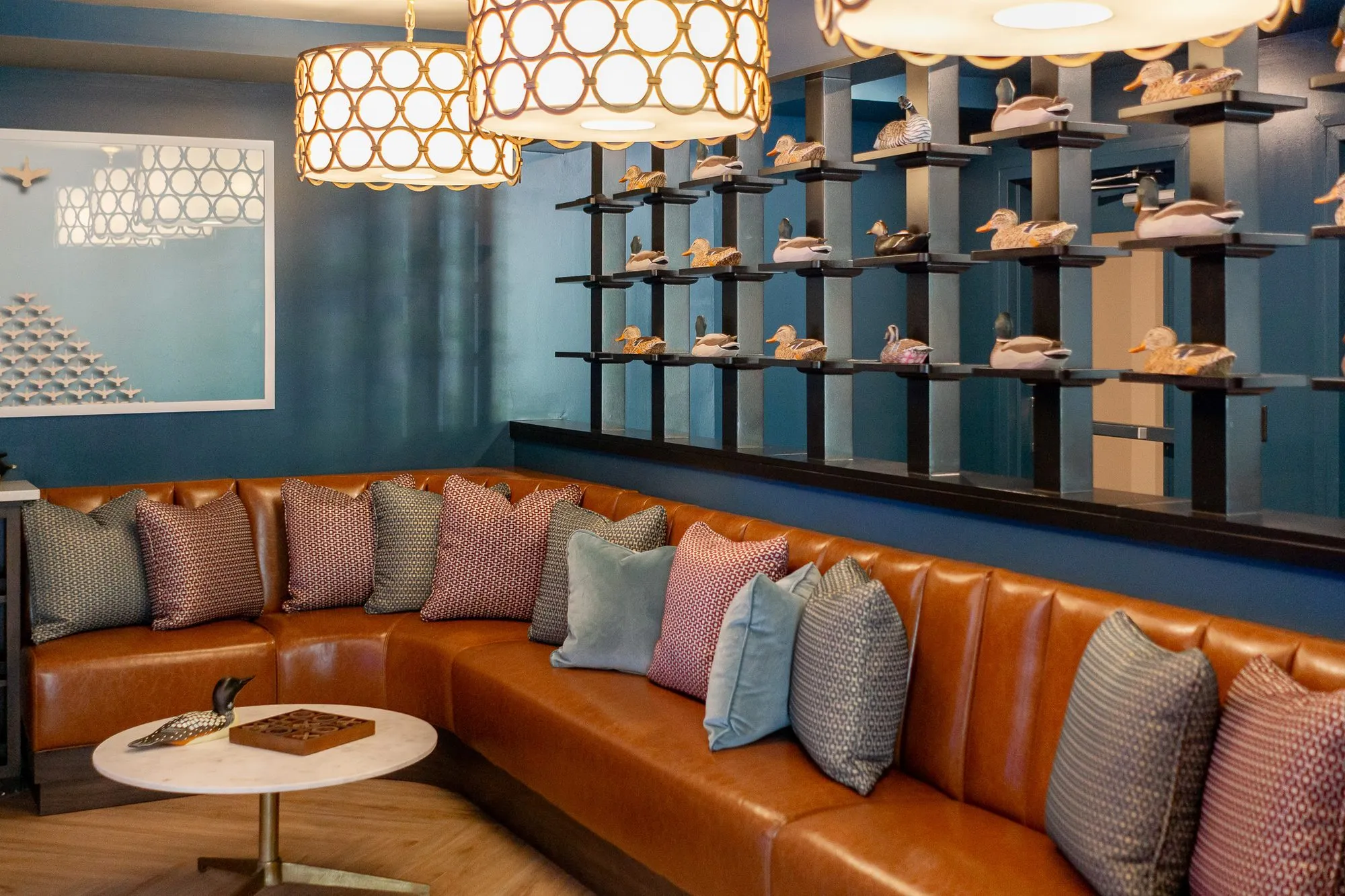 Multifamily amenity bowling lounge with a cognac leather corner banquette under ring-pattern pendants, duck decoys above
