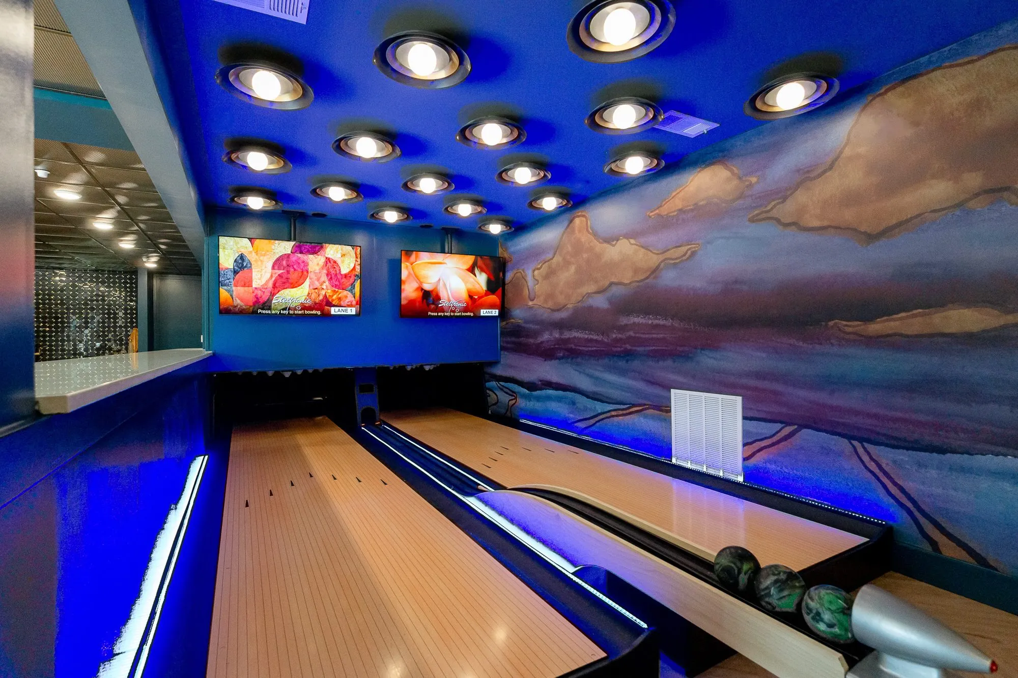 Multifamily amenity two-lane duckpin bowling alley under blue light, a painted landscape mural and screens at the end