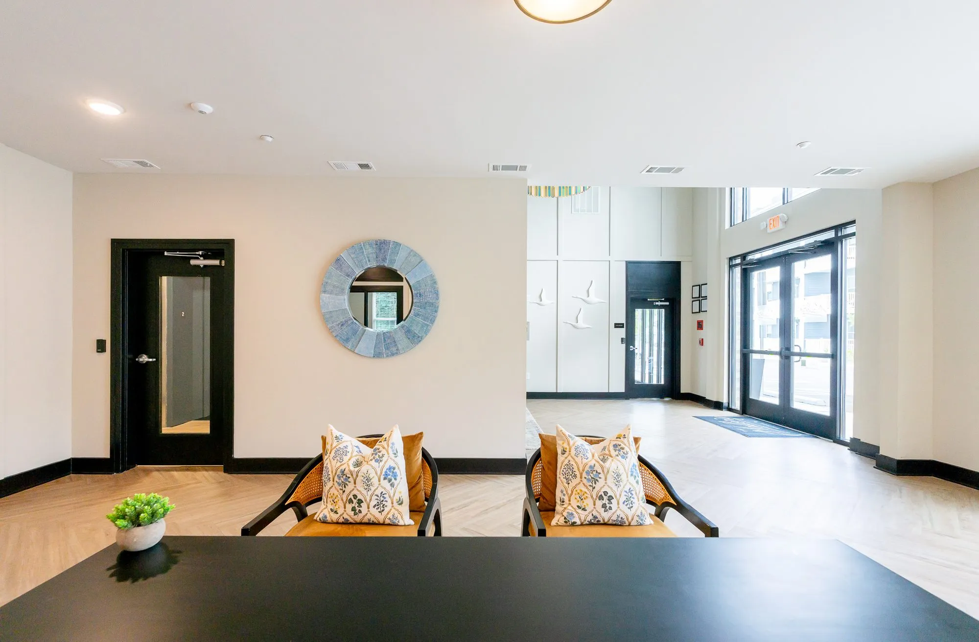Multifamily amenity leasing area over a black table to two cane chairs before a round mirror, glass entry doors at right