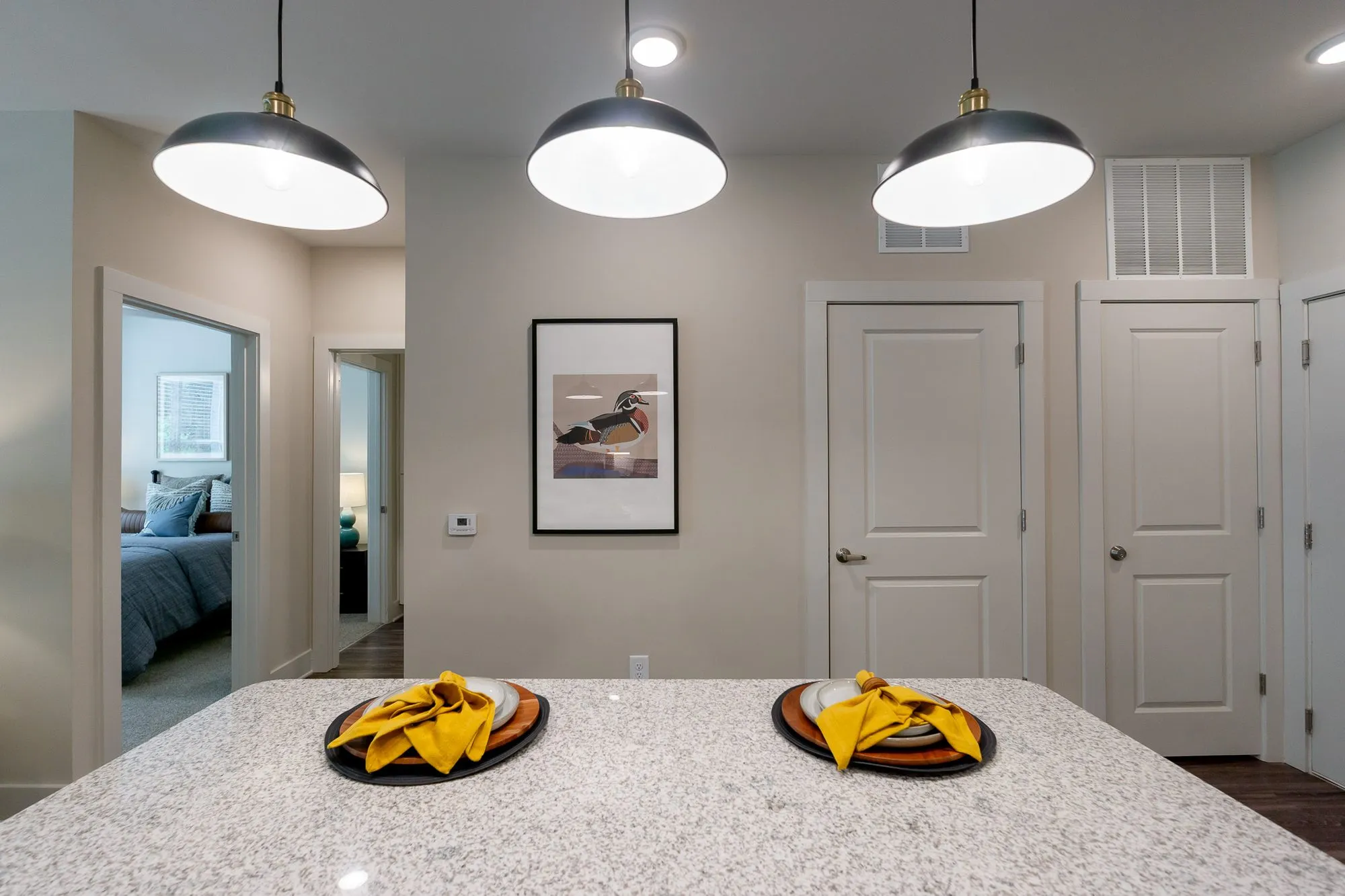 Multifamily apartment model kitchen island set with yellow napkins under black dome pendants, a bedroom beyond