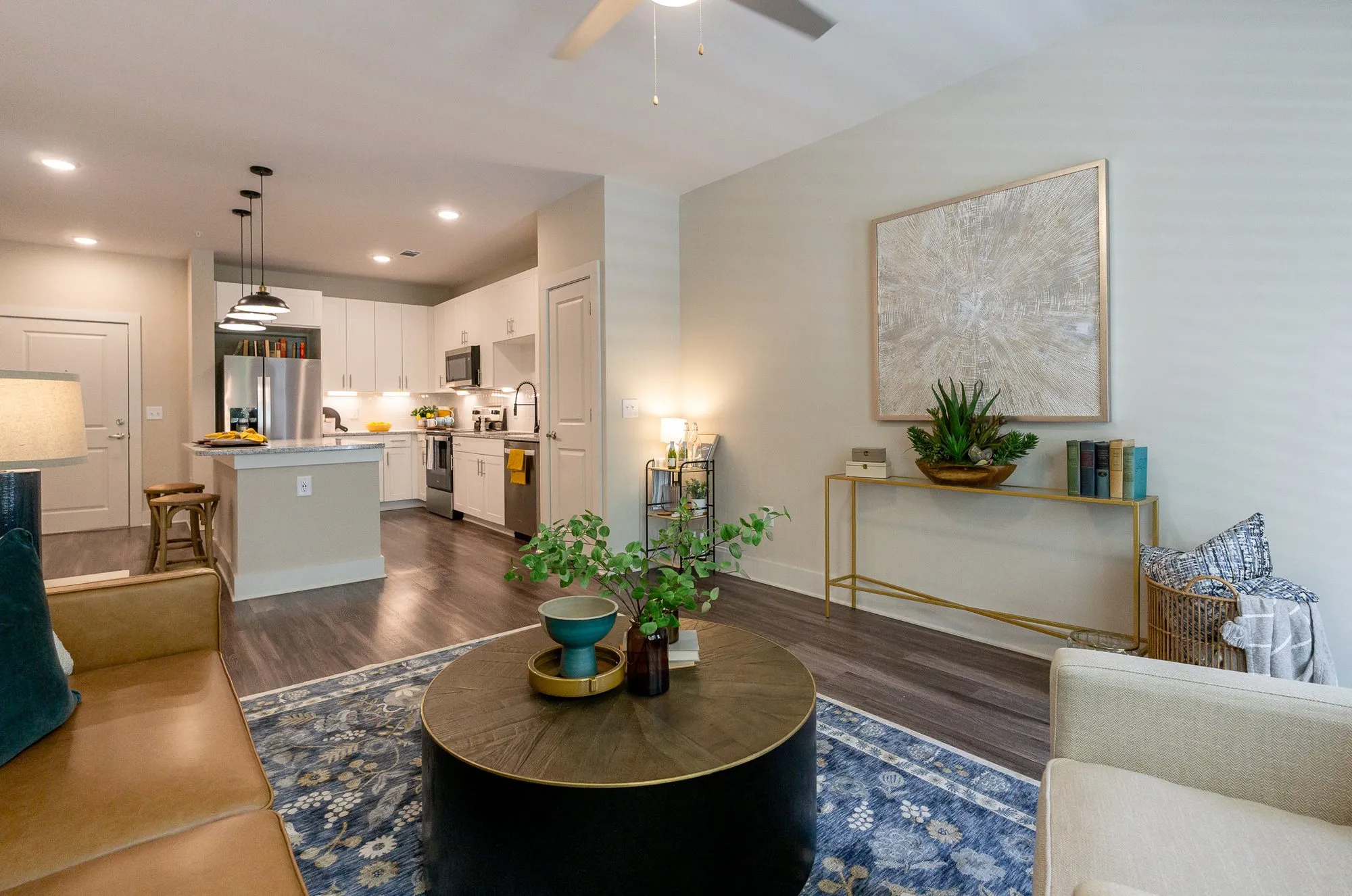 Multifamily apartment model living area with a cognac sofa and round coffee table on a blue rug, the kitchen beyond